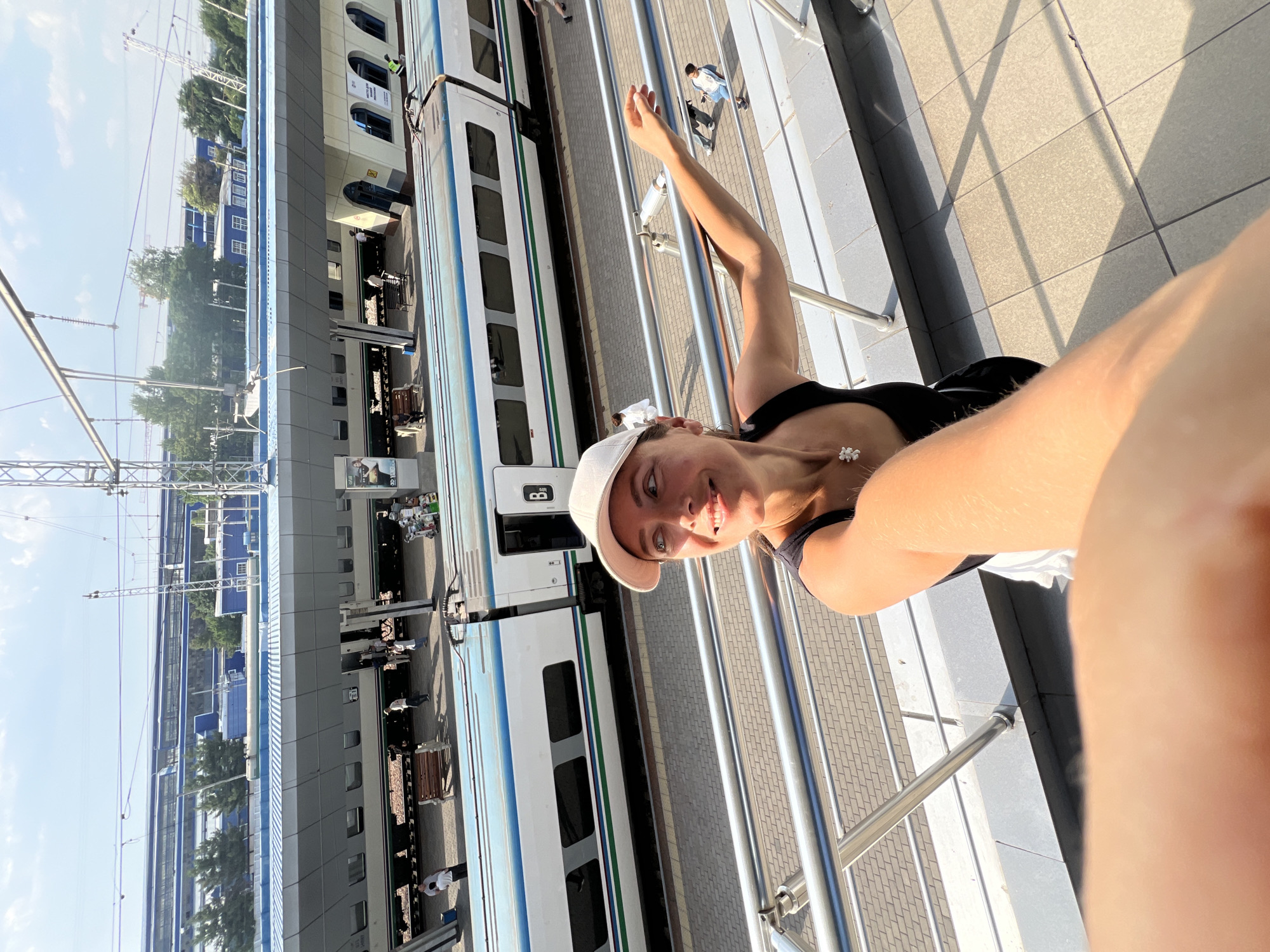 A woman wearing a black tank top and a light-colored baseball cap takes a selfie at a train station. She is smiling and looking to her left. Her arm is extended forward, holding the camera. A train is visible in the background, as are several people on the platform. The train station appears modern with blue accents on the building and the train itself has blue and green stripes. The pavement is tiled. The scene is brightly lit by sunlight, casting shadows. The overall setting suggests an outdoor urban environment, likely a public transportation hub. The image includes details that suggest the location is Tashkent, Uzbekistan, such as the architectural style of the station and the design of the train.