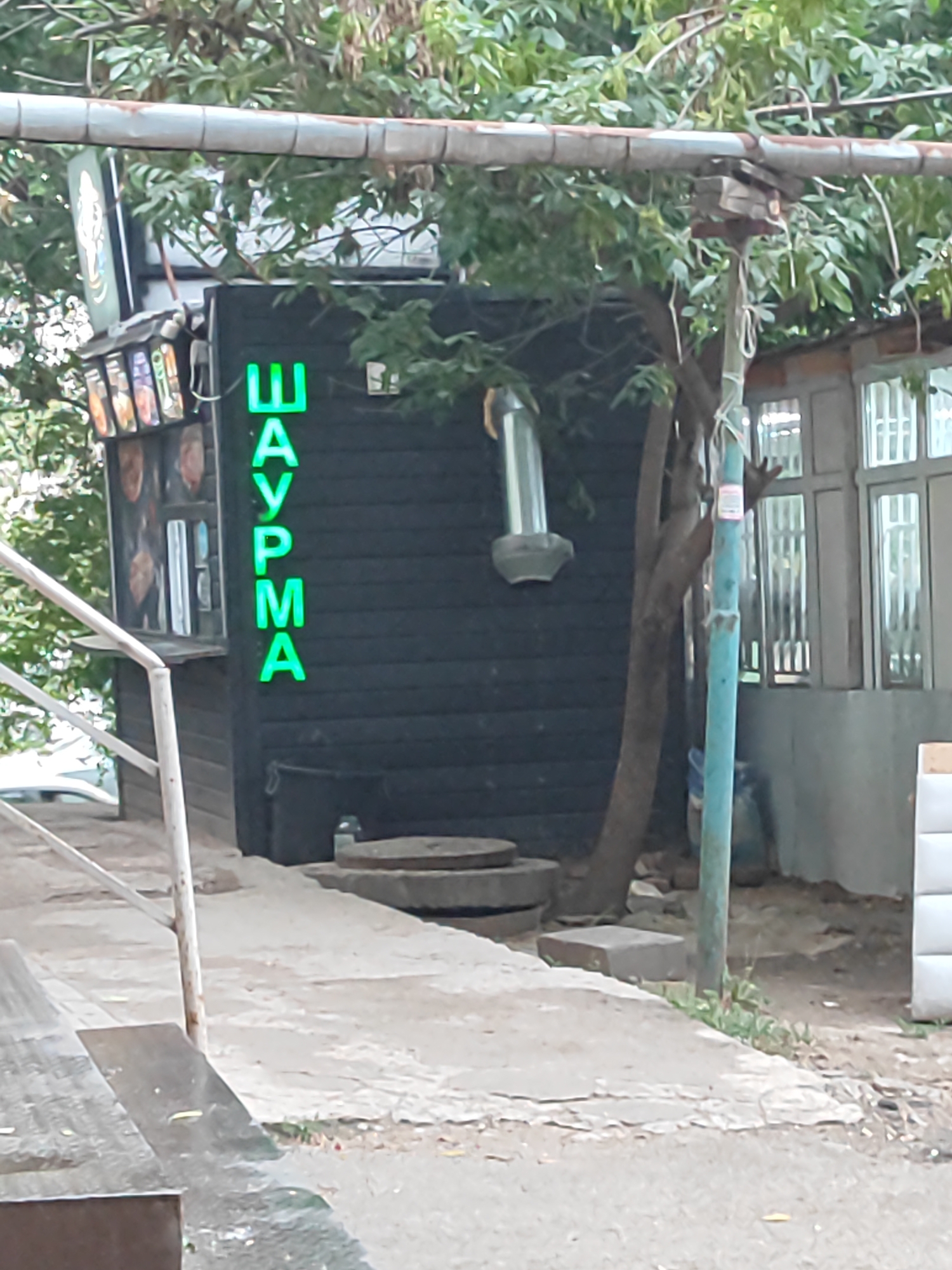 The image shows a street-level view of a small, dark-colored food kiosk with bright green neon lettering spelling "ШАУРМА" (SHAURMA) vertically on its facade. The kiosk appears to be located outdoors, with trees and foliage visible in the background. A metal pipe runs horizontally above the kiosk, supported by posts. To the left of the kiosk, there is a window displaying pictures of food items. To the right, a light blue pipe stands vertically, and a building with windows is visible. The ground in front of the kiosk is paved with concrete, and there are some circular concrete structures and a small brick on the ground. The scene suggests an urban environment, likely a street or sidewalk, in Almaty, Kazakhstan, given the context. No people are visible in the image.