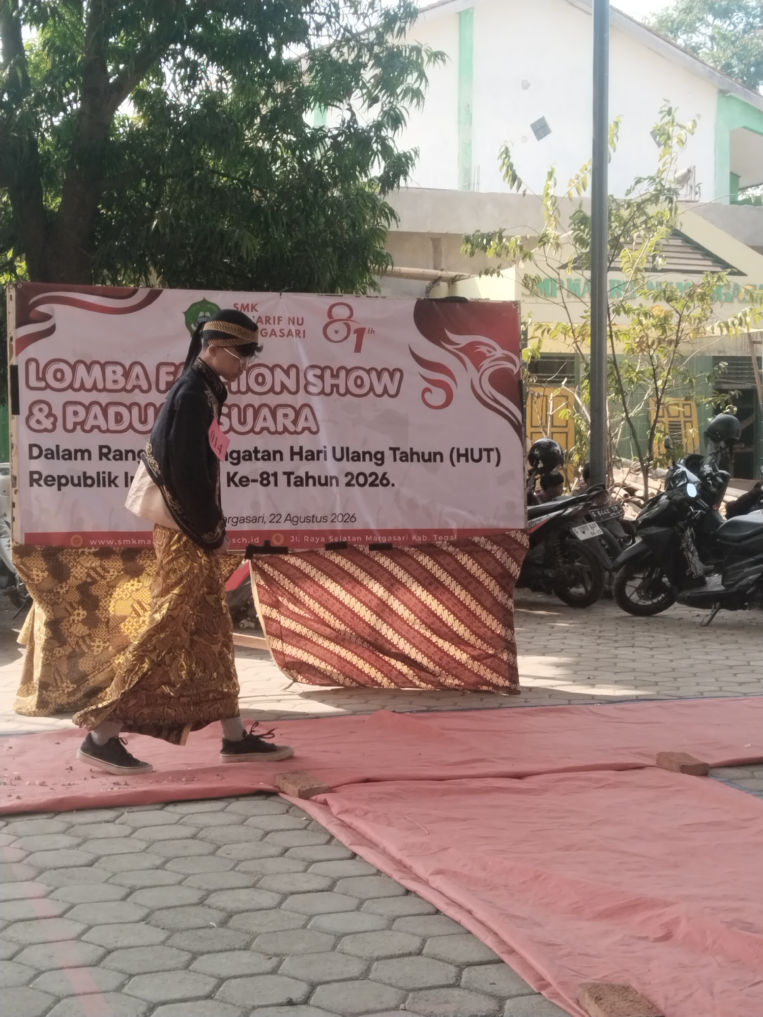 A person, dressed in traditional Indonesian attire consisting of a dark, long-sleeved jacket, a white undergarment, and a patterned golden sarong, walks on a pink tarp laid over a hexagonal paved surface. The person wears glasses and a black head covering with a decorative band. They are walking on what appears to be a makeshift runway. Behind the person is a large banner with red and white lettering announcing "LOMBA FASHION SHOW & PADU SUARA" and details about the 81st anniversary of the Republic of Indonesia. The banner also includes website and address information: "www.smkma...", "sch.id", and "Jl. Raya Selatan Magasari, Kab. Tegal." To the right of the banner, several motorcycles are parked. The background shows a building with a white and green facade and trees. The scene is outdoors, likely in a school or public area in Pakulaut, Indonesia.