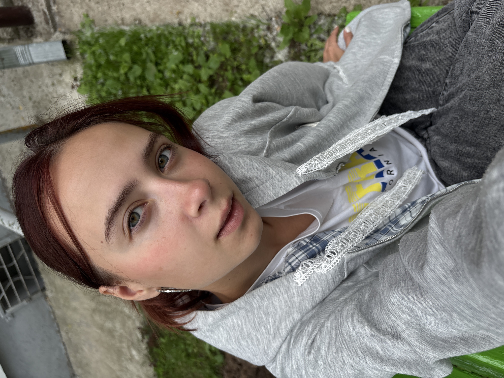 The image shows a young woman with auburn hair and green eyes, wearing a gray zippered hoodie over a plaid shirt and a white t-shirt. She has a silver hoop earring in her left ear. She is sitting on a green bench, outdoors, with a patch of green grass and a concrete walkway behind her. A metal drainpipe is visible on a building in the background. The overall scene appears to be in an urban or semi-urban setting, possibly a courtyard or park area. The specific location in Chaikovsky, Russia, is not explicitly identifiable from the image alone.