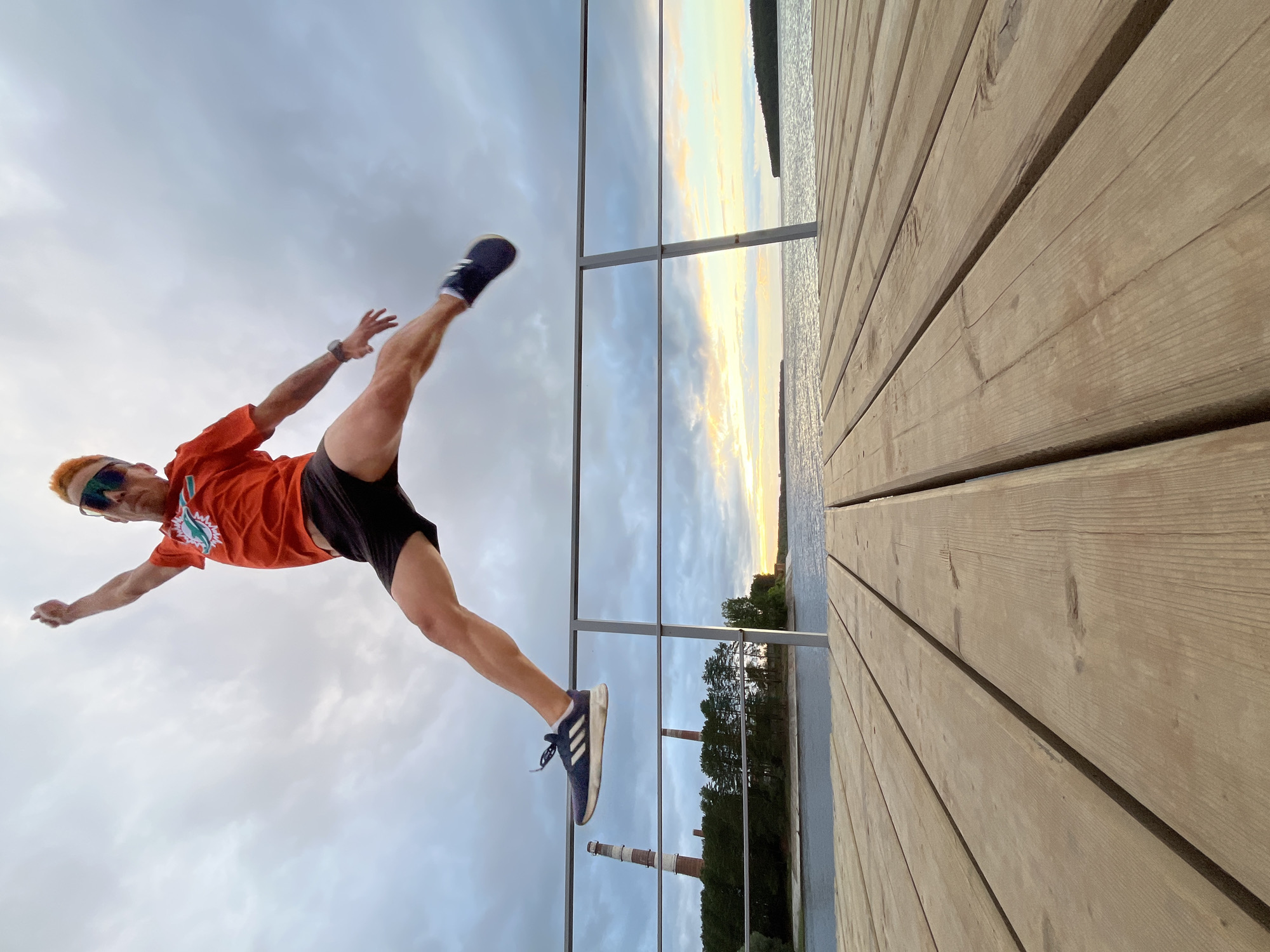 A man with orange hair, wearing an orange t-shirt, black shorts, and blue and white athletic shoes, is captured in mid-air, performing a dynamic kick. He appears to be on a wooden pier or boardwalk, with a metal railing to his left. The pier's wooden planks are prominent in the foreground, running diagonally across the frame.
Behind the railing, a body of water stretches into the distance, reflecting the sky. The sky itself is a mix of cloudy grey and patches of warm, soft light, suggesting either sunrise or sunset. The horizon is lined with trees and two tall, striped industrial chimneys, indicating an urban or industrial setting within a natural landscape. The water and sky create a serene, diffused backdrop.
The man's pose, with one leg extended high and arms outstretched, conveys a sense of energy, athleticism, and perhaps playfulness or freedom. The lighting suggests it is either early morning or late evening. The overall scene is one of outdoor activity, possibly exercise or a moment of joyful expression, set against a quiet, natural environment with a hint of industrial presence. The logo on the man's shirt is partially visible but not clearly legible.