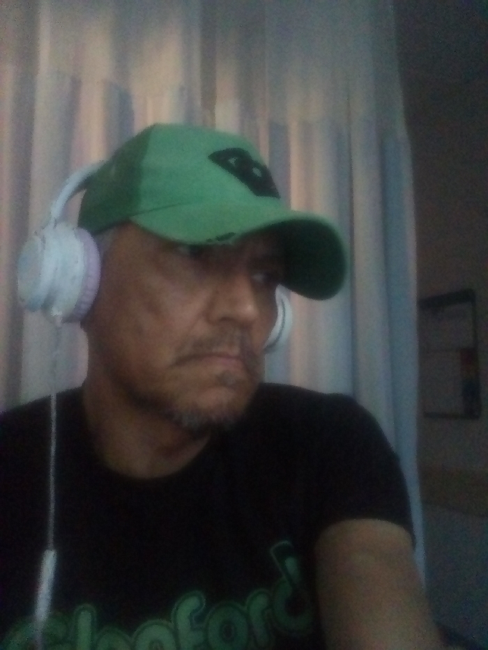 A man in a black t-shirt and green baseball cap is wearing white headphones. He has short gray hair and a goatee. His expression is serious. The background features striped curtains and a whiteboard. The lighting is subdued, and the image quality is somewhat grainy.