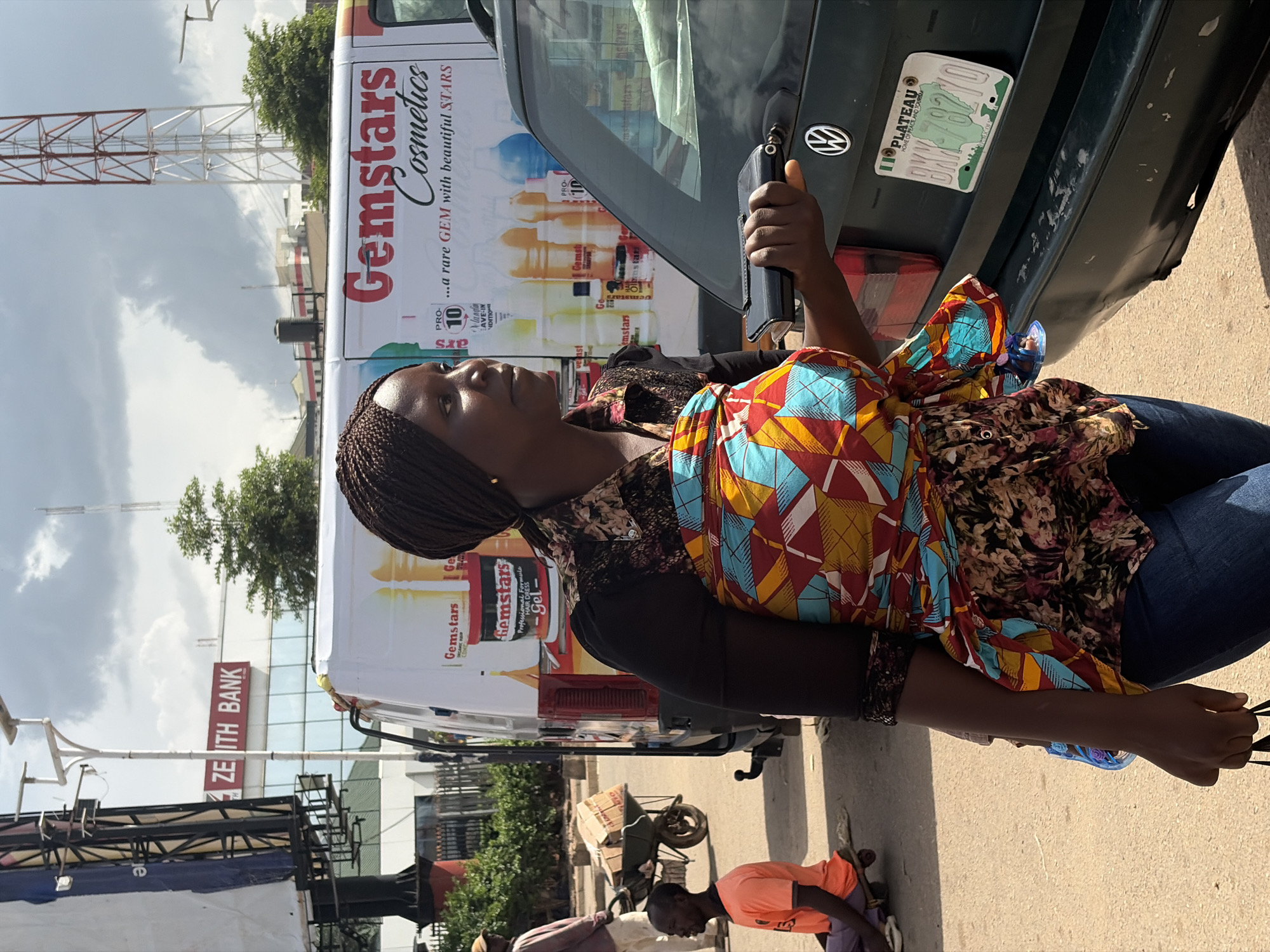 This media file shows a woman with braided hair, wearing a colorful patterned cloth tied around her waist. She is standing next to a vehicle, with a partially visible license plate that reads "PLATEAU BXY-78270 HOME OF PEACE AND TOURISM." The side of the vehicle features advertisements for "Gemstars Cosmetics" and "Gemstars Professional Formula HAIR DRESS -Gel-". In the background, there are buildings with signage for "ZENITH BANK," a communication tower, and trees. Other people and a wheelbarrow are visible in the lower part of the image, suggesting an outdoor street scene. The lighting suggests it is daytime, and the sky appears cloudy. The overall impression is a candid, everyday moment captured in a commercial or urban setting in Jos, Nigeria.