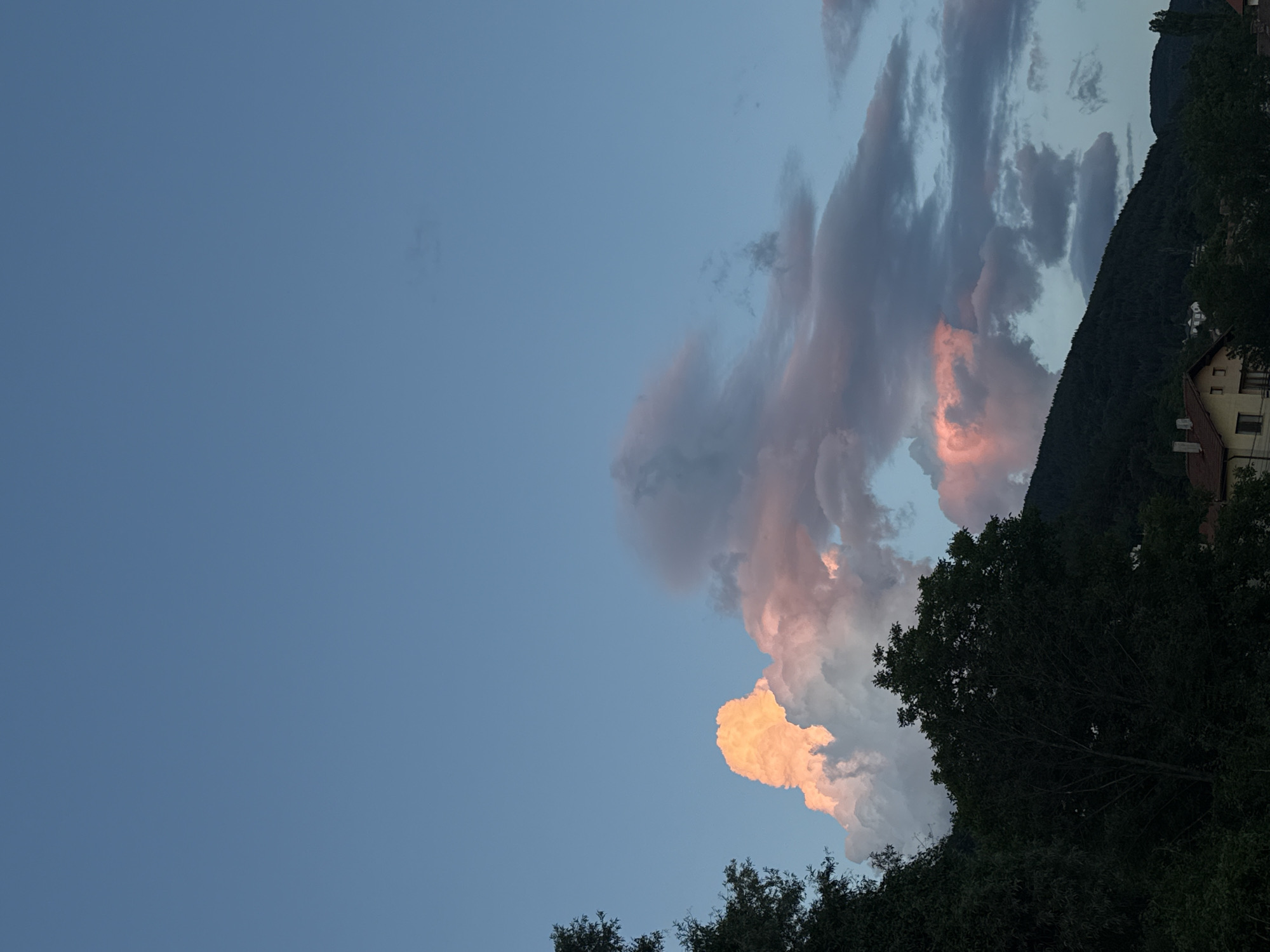 This media file depicts a twilight scene of clouds illuminated by the setting sun over a mountainous landscape. The sky is a gradient of light blue, transitioning to darker hues near the horizon. Dramatic clouds, tinged with vibrant oranges and pinks, dominate the right side of the frame, suggesting the last rays of sunlight catching their edges.
The background features a dark, tree-covered mountainside, on which a building with red-tiled roofs is partially visible. In the foreground, the silhouettes of trees are prominent, their dark foliage contrasting with the illuminated sky.
The overall mood is serene and picturesque, characteristic of a peaceful evening. The time of day is clearly sunset or twilight, given the colors of the clouds and the low angle of the sun. The location cues point to a mountainous region, possibly a village nestled in the foothills, as suggested by the building and the steep terrain.
No people or specific objects are clearly discernible, beyond the natural elements of sky, clouds, trees, mountains, and the building. There is no visible text in the media file.