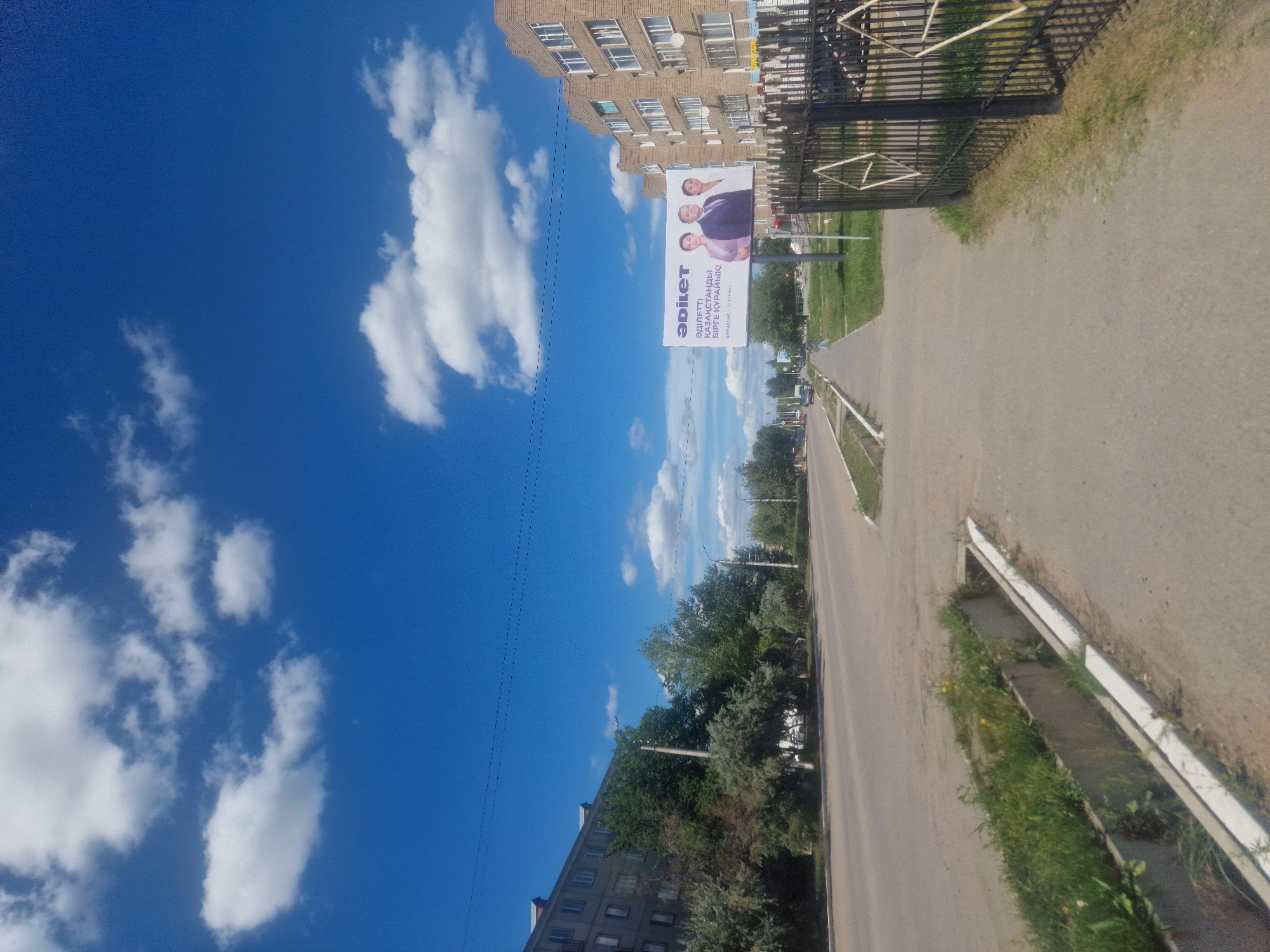 A wide, paved road runs through an urban environment. On the left side of the road are several apartment buildings and a line of trees. On the right side of the road, there is a sidewalk, a black metal fence, and a billboard with three people pictured on it. The billboard has text in a foreign language that appears to be Kazakh. The sky above is a vibrant blue with scattered white clouds. The overall impression is of a sunny day in a small city or town.