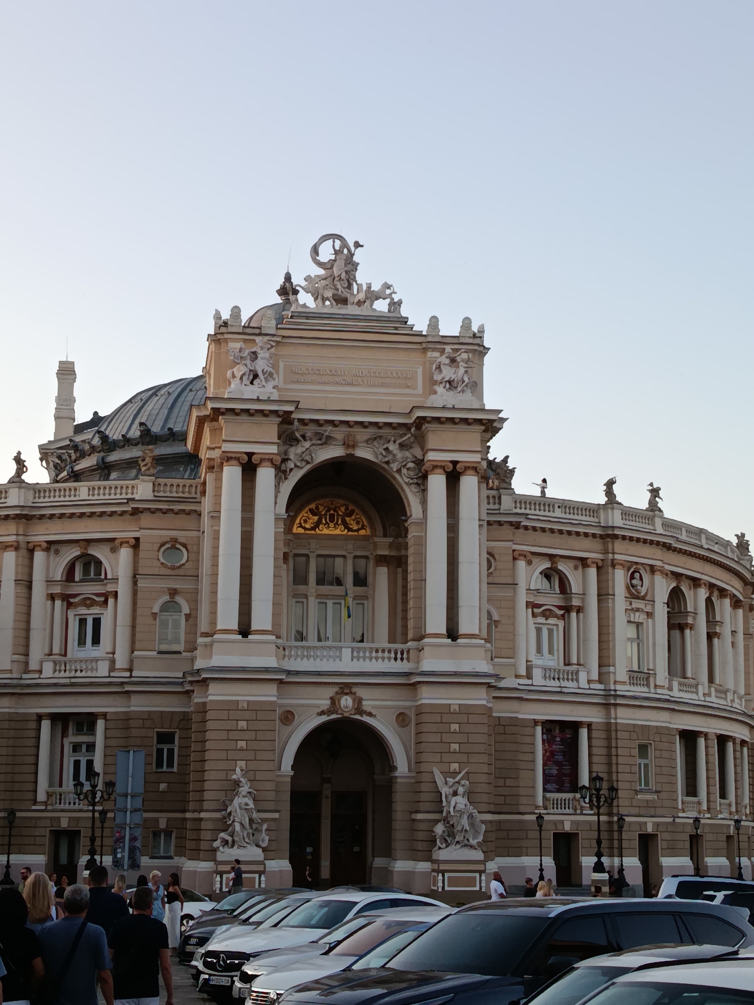 The image shows the exterior of a grand, ornate opera house, likely in a European city, with a clear sky above. The building features classical architecture with columns, sculptures, and decorative elements. In the foreground, several cars are parked, and a group of people are walking or standing, some looking towards the building. The overall scene suggests a public gathering or a visit to a cultural landmark. The architecture and style of the building could be indicative of cities like Odessa, Vienna, or Prague, but without further information, the specific location remains undetermined.