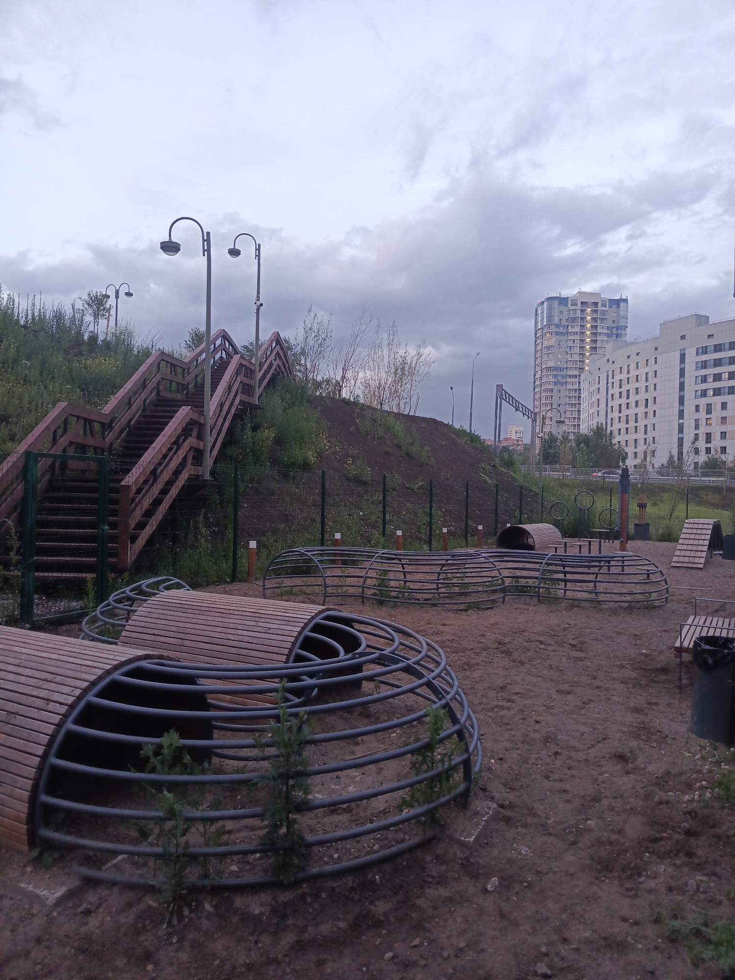 The image depicts an outdoor dog park in Perm, Russia, under an overcast sky. In the foreground, there are several semi-circular wooden structures with metal framing, designed as agility obstacles for dogs. The ground is covered with sand and dirt.
Behind the dog park, a large wooden staircase ascends a grassy embankment. Several lampposts are visible along the walkway leading to and from the staircase. In the background, modern residential buildings rise above the trees, with a slightly hazy atmosphere. The overall scene suggests a public recreational space in an urban environment. There are no people or animals visible in the image.