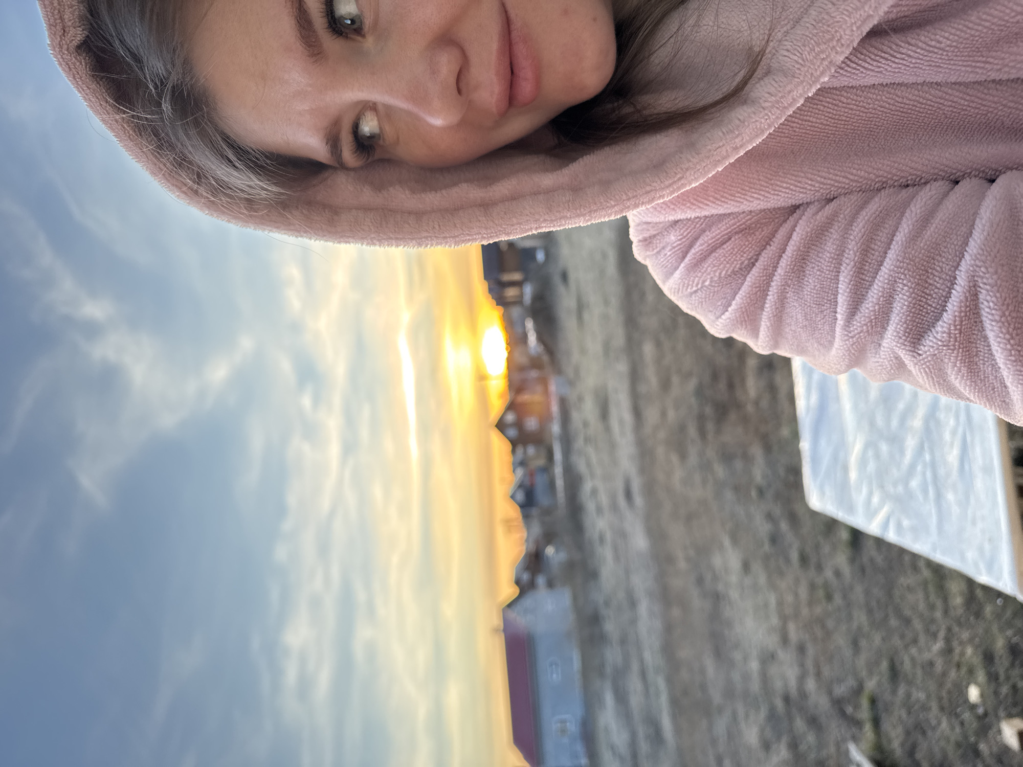 The image, rotated 90 degrees counter-clockwise, presents a close-up selfie of a woman against a picturesque sunrise or sunset. The woman occupies the upper half of the frame, looking towards the camera with a serene, slight smile. Her light-colored eyes, nose, and lips are clearly visible. She wears a soft, pink, hooded garment, possibly a terry cloth robe or sweatshirt, with the hood drawn around her head.
The background showcases a vibrant sky transitioning from a soft blue on the left to a radiant golden-orange on the right, where the sun is either rising or setting, casting a warm glow. Sparse, wispy clouds are scattered across the sky. Below the horizon, a desolate landscape of what appears to be dry, low-lying ground or sparse vegetation stretches into the distance. Several small, indistinct buildings are visible on the horizon line, suggesting a rural or suburban area, consistent with the location context of Gornyi (Dvurechenskoe S/P), Russia. On the far right, near the bottom, a portion of a translucent white object, possibly a plastic sheet, is partially visible. The overall mood of the image is calm and reflective, capturing a peaceful moment during the golden hour. No text is visible.