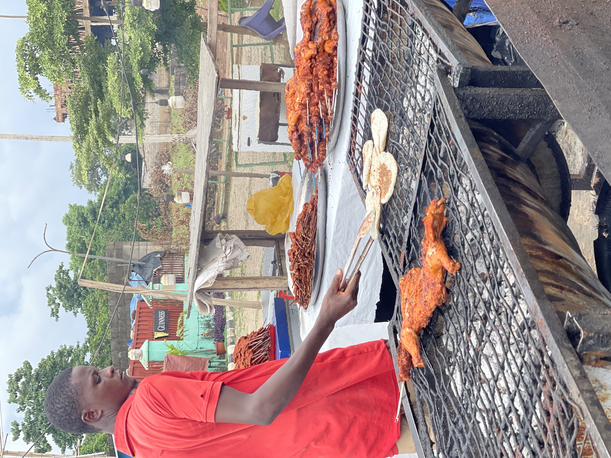 A young man in a red polo shirt is grilling chicken at an outdoor food stall in Galadimawa Village, Nigeria. The chicken pieces are coated in a reddish-orange marinade and are arranged on a metal grill over coals. Beside the grill are platters of grilled chicken skewers and other fried snacks. In the background, there are trees, buildings under construction, and a sign that says "Guinness." The scene appears to be a typical roadside food vendor operation.