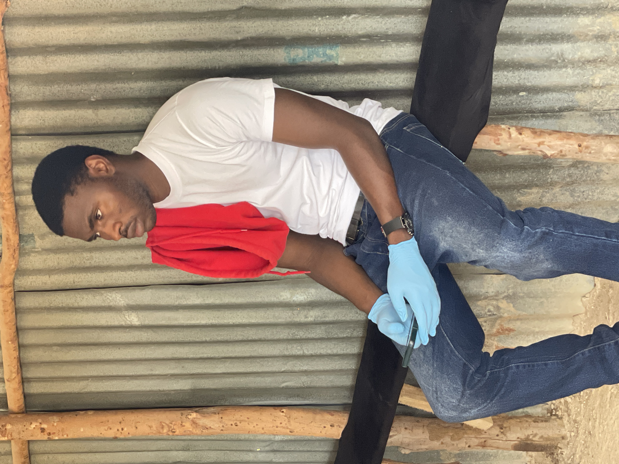 A young Black man is pictured from the chest down, angled slightly to his left, looking down. He is wearing a white t-shirt, blue jeans, a black watch on his left wrist, and light blue disposable gloves on both hands. He is holding a dark, slender object, possibly a pen or stylus, in his gloved right hand, with his left hand resting on a bright red piece of fabric. The background consists of corrugated metal walls and wooden beams, suggesting an unfinished or rustic structure. The lighting appears to be natural, indicating daytime. The man's facial expression is neutral and focused. The setting is likely outdoors or in an open-air structure given the materials used for the walls and supports. There is no visible text.