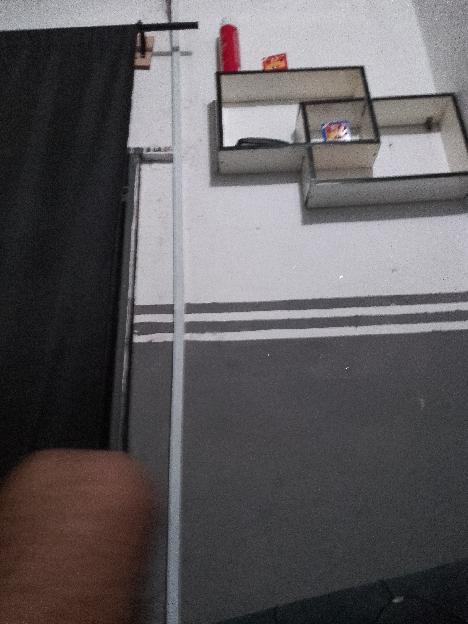 The image shows a close-up of a wall with a dark curtain on the left side, a white vertical bar, and a three-tiered shelf unit. The shelf unit is mounted on the wall and contains a red aerosol can, two small rectangular boxes, and what appears to be a black electronic device. The wall has two horizontal stripes painted in grey and white. The lower portion of the image shows a blurred brown object, possibly a person's arm or leg. The scene appears to be indoors, in a residential setting. The image is grainy and slightly out of focus. There is no indication of the specific location within Jauro Yinu, Nigeria.