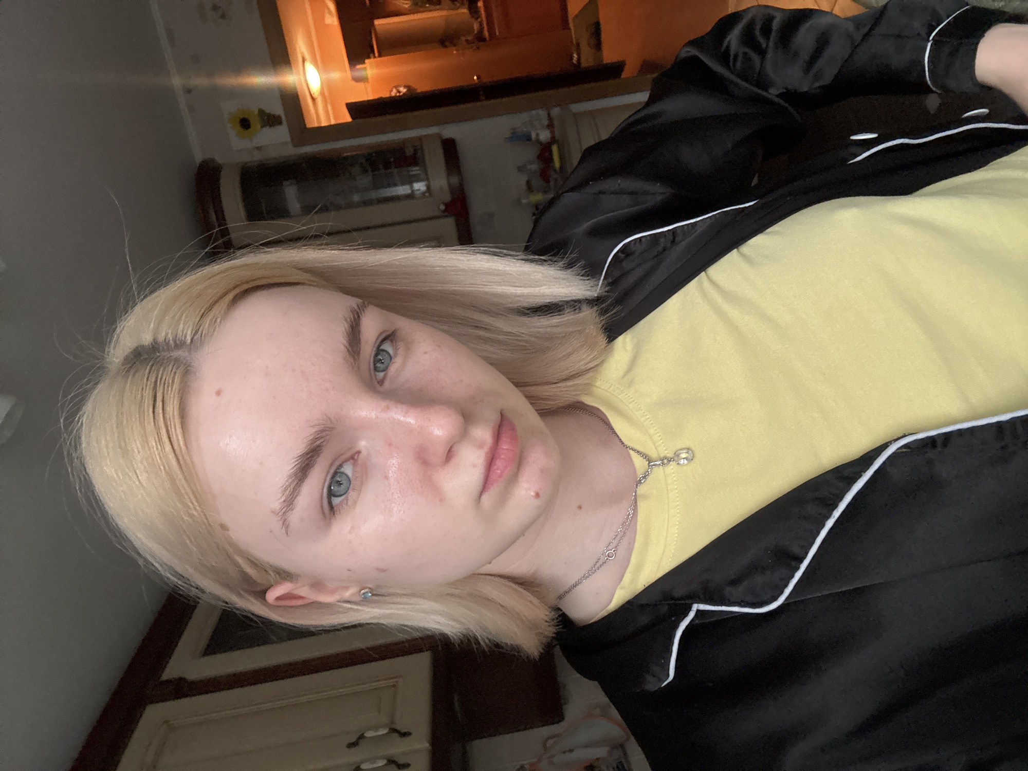The image shows a close-up, waist-up, indoor selfie of a young woman with blonde hair and blue eyes. She is wearing a yellow t-shirt and a black pajama-style robe with white piping. A silver necklace with a pendant is visible. She has light skin with some freckles and a few blemishes. The background appears to be a kitchen, with wooden cabinets and a doorway leading to another room. The lighting suggests it is daytime, with natural light coming from a window or overhead fixture, and a warm artificial light source visible in the background. The specific location within Sinyavino, Russia cannot be determined from the visual information provided.