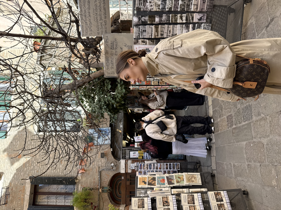 The scene depicts a narrow, cobbled pedestrian alleyway in Venice, Italy, flanked by aged brick buildings under natural daylight. In the right foreground, a young adult female, with light brown hair pulled back, stands in profile facing left. She wears a tan, high-collared trench coat and carries a round, brown Louis Vuitton monogrammed crossbody bag. Her right hand, resting on the bag, holds a dark object, likely a mobile phone.
In the background, several individuals are visible: a person with dark red hair wearing a black top and a long white pleated skirt, and a second person in a light-colored oversized jacket and dark pants, carrying a black shoulder bag, both facing away from the camera. Further back, another individual in a tan top and dark pants is visible, looking at a phone.
The alley walls feature a dark wooden arched doorway on the left, an upper-floor window with dark green shutters, and a wall-mounted lantern. A large, leafless tree with intricate branches dominates the upper center, with green foliage below it. Metal display racks filled with postcards, calendars, and books line both sides of the alley. Signs affixed to the wall are visible, including one in Italian and English stating, "WE KINDLY INFORM OUR VISITORS THAT SCHOOL AND SCHOOL GROUPS CONSISTING OF MORE THAN 10 PEOPLE ARE REQUIRED TO BOOK," and another in Italian requesting visitors to hold their backpacks. The ground is paved with irregular stone blocks.