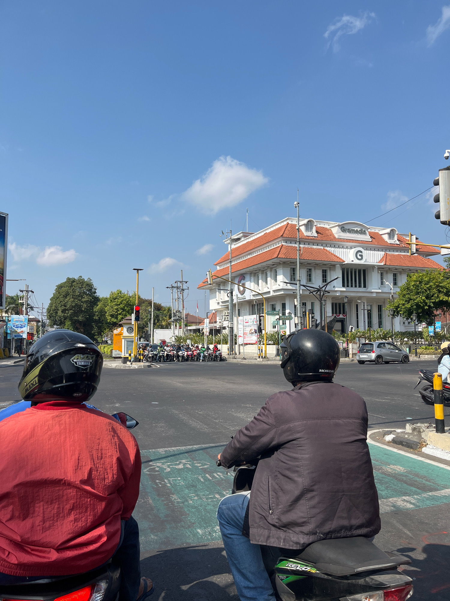 This is a street scene in Yogyakarta, Indonesia, likely at a busy intersection. Two individuals are seated on motorcycles, viewed from behind, awaiting the traffic signal. The person in the foreground on the left wears a red jacket and a black helmet with yellow accents. The person on the right wears a dark jacket and a black helmet. The motorcycles are stopped at what appears to be a pedestrian crossing marked with green paint.
In the background, a prominent white building with a red tiled roof stands out. It features architectural details like arched windows and ornate cornices. The word "Gramedia" is visible in large letters on the facade, indicating it is a bookstore or media center. Several traffic lights are present, with one showing a red signal. A line of motorcycles is stopped at the intersection, also waiting for the light. Trees and utility poles line the street. The sky is clear and blue with a few scattered clouds, suggesting a sunny day. The overall impression is a typical urban street scene in a bustling Indonesian city.