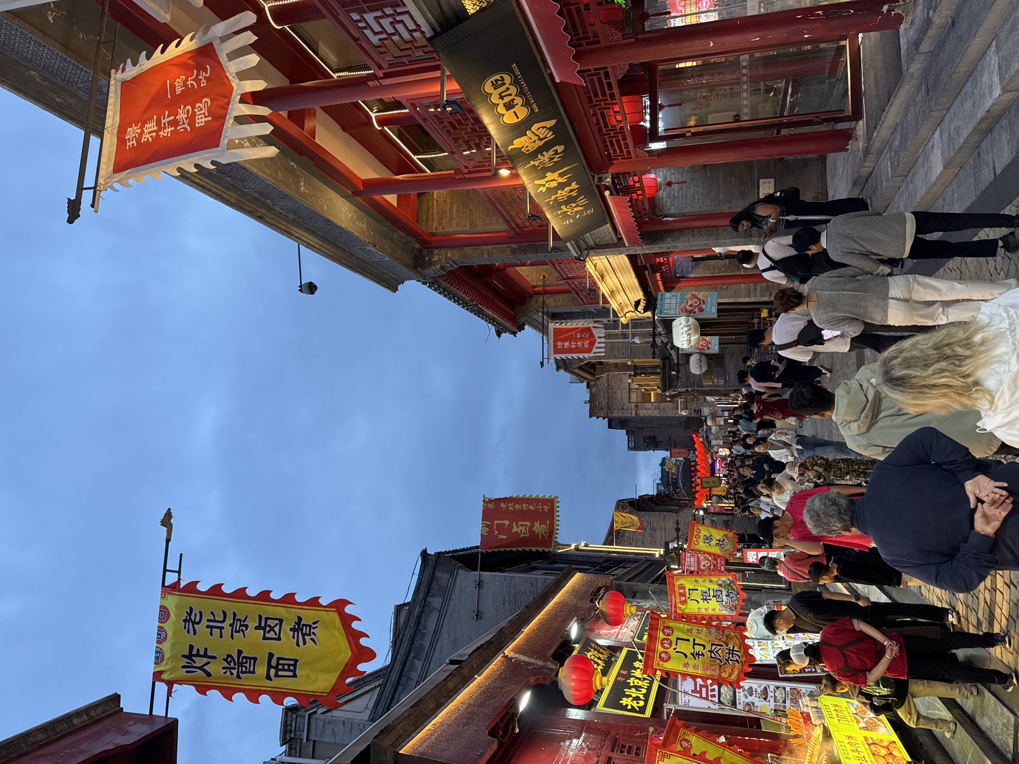 The image captures a bustling street scene in Dongcheng District, China, likely a popular tourist or food area. A dense crowd of people meanders along the street, primarily looking at or interacting with the numerous food stalls lining both sides. The architecture prominently features traditional Chinese elements, with red pillars, ornate roof structures, and hanging lanterns. Many signs with Chinese characters advertise different foods and services, contributing to the vibrant and colorful atmosphere. The time of day appears to be dusk or early evening, as the sky has a blue hue and some lights are already illuminating the scene. The focus of activity is clearly on the food and the experience of exploring the local culture.