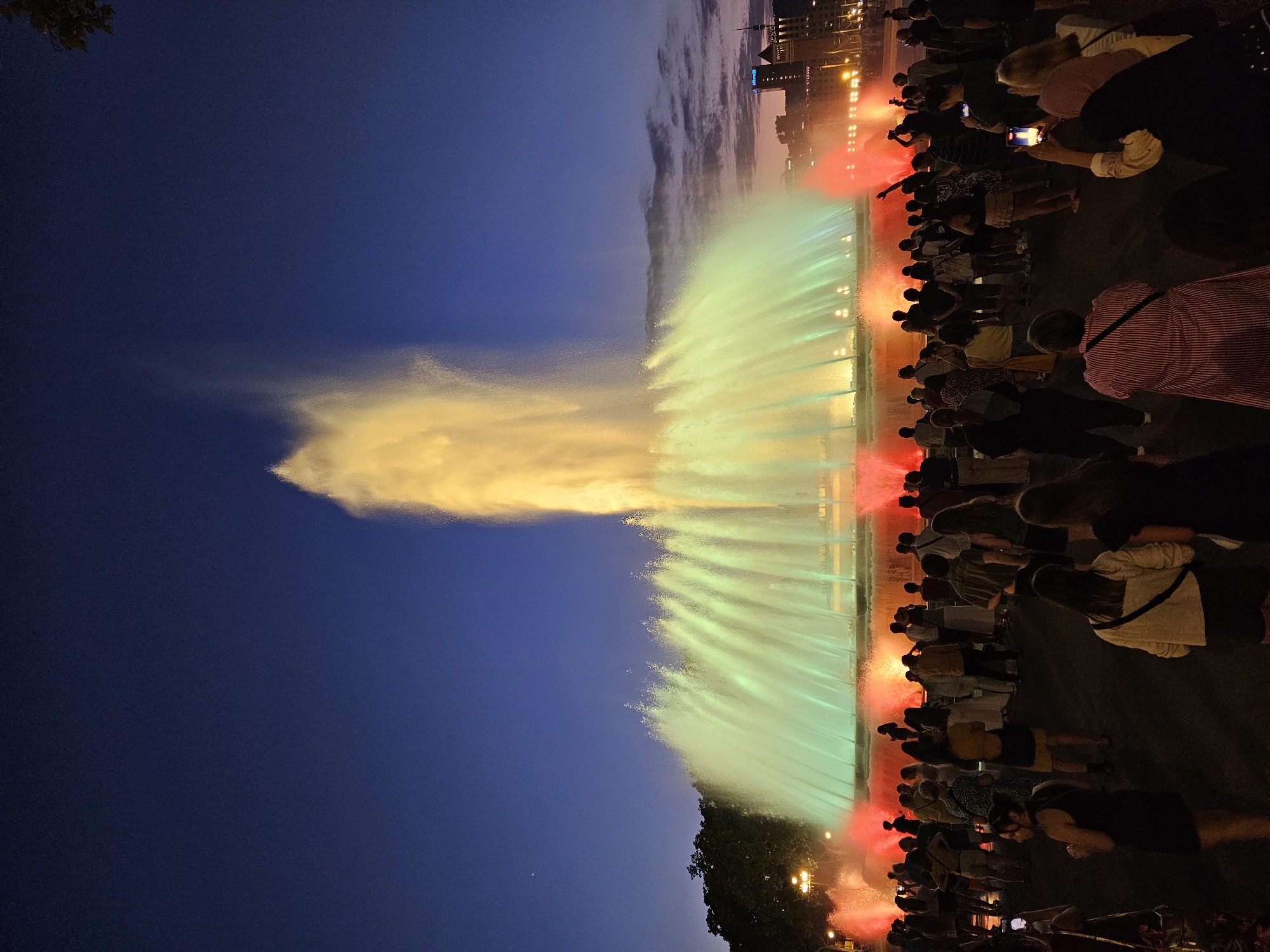 This image captures a vibrant nighttime scene at the Magic Fountain of Montjuïc in Barcelona, Spain. A large, illuminated fountain is the central focus, with plumes of water shooting upwards and outwards, lit by a dynamic array of colors. The water spray is predominantly illuminated with a bright yellow light at the top, transitioning to vibrant green and red hues lower down, creating a dramatic and colorful spectacle against the dusky twilight sky.
The fountain is surrounded by a substantial crowd of people, gathered to watch the display. They are silhouetted against the bright lights of the fountain and the city, with some individuals captured holding up phones, presumably to record or photograph the event. The crowd is dense in some areas, indicating a popular attraction.
In the background, the Barcelona cityscape is visible, with buildings and streetlights suggesting an urban environment. The sky transitions from a deep blue at the top to lighter shades of purple and pink closer to the horizon, with some clouds visible, hinting at the fading light of dusk. Trees and lampposts are also present in the mid-ground, adding to the atmosphere of an outdoor public space. The overall impression is one of a lively evening gathering, with the Magic Fountain providing a spectacular visual performance for the assembled spectators.