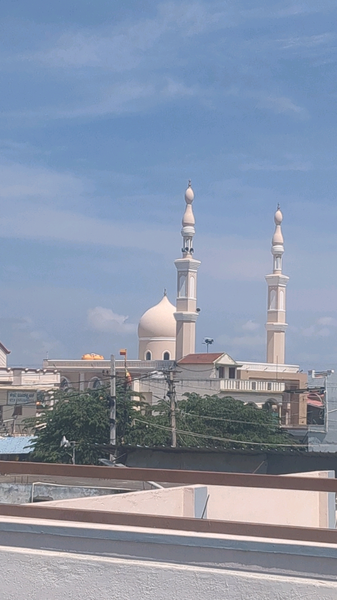 The image depicts a mosque with two minarets and a dome, set against a bright blue sky with scattered clouds. The mosque appears to be a prominent structure in the city of Alipur, India. In the foreground, there are rooftops and electrical wires, suggesting an urban environment. The architecture of the mosque features light-colored walls and intricate detailing on the minarets and dome. The surrounding buildings are visible, showcasing a mix of architectural styles. The overall scene suggests a clear and sunny day, with the mosque standing as a significant landmark in the cityscape.