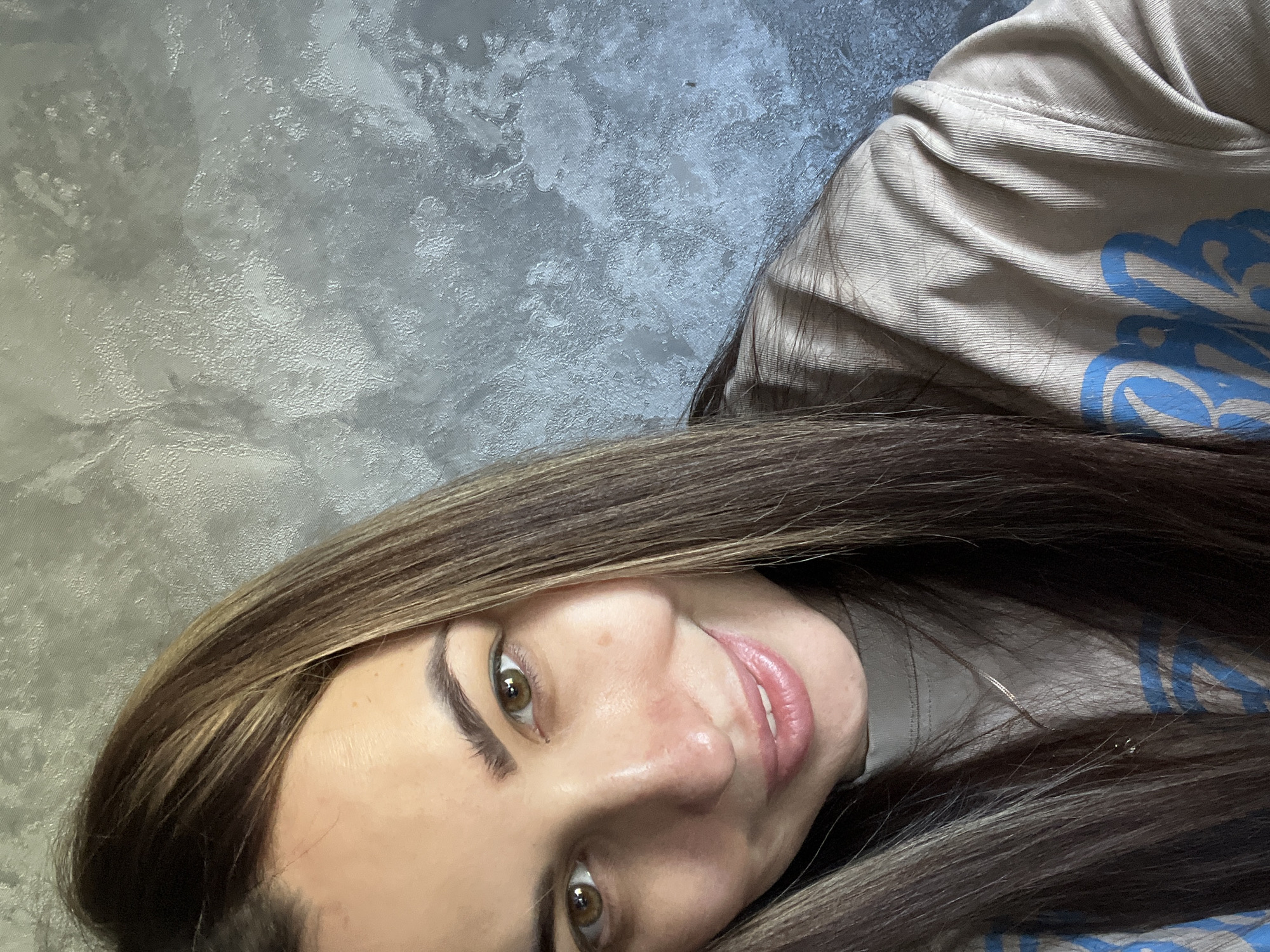 A selfie of a woman is oriented with the upper left corner as the top. The woman has long, light brown hair with highlights. She is wearing a beige sweatshirt with blue lettering. Her eyes are hazel, and she is smiling slightly. The background is a textured grey wall. The image does not contain information regarding location.
