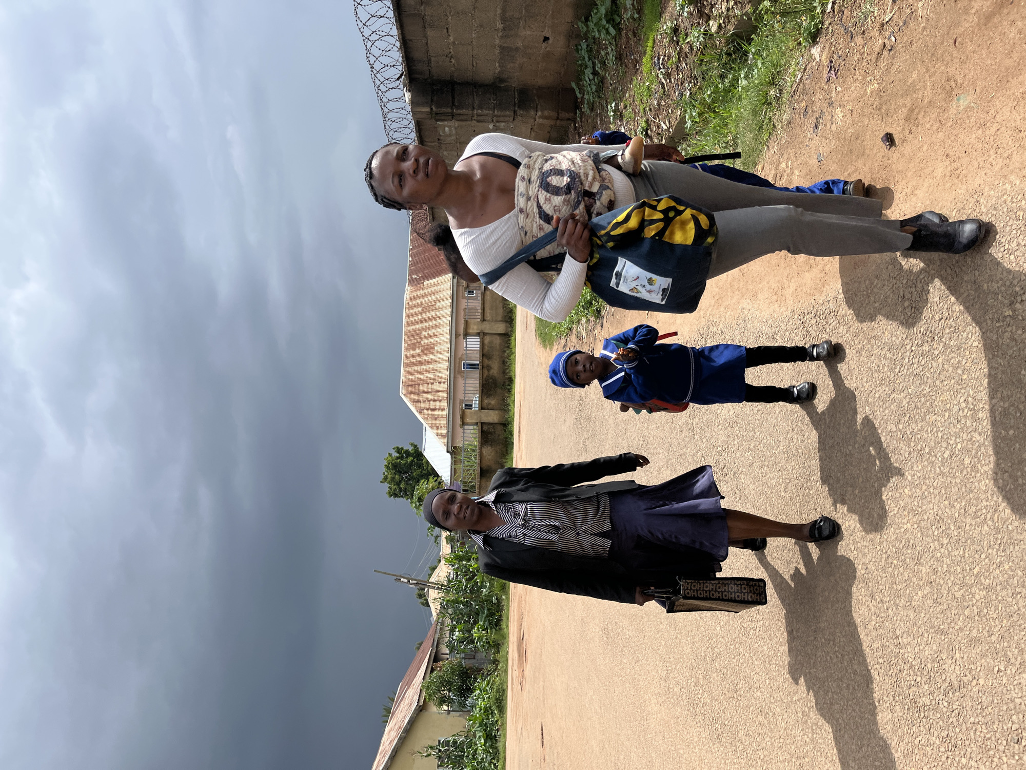 In Jos, Nigeria, under a stormy, dark grey sky, two women and two children walk along a dirt road. The woman on the left wears a black blazer over a patterned shirt and a dark blue skirt. She carries a large black tote bag. The woman on the right, holding a child strapped to her back, wears a white long-sleeved shirt and grey pants. She has a large blue and yellow bag slung over her shoulder. A small child in a blue uniform and hat walks in front of the woman on the right, holding their hands together. Buildings with corrugated metal roofs are visible in the background, along with a fence topped with barbed wire. The ground is a dusty brown, with shadows cast by the people and the uneven terrain.