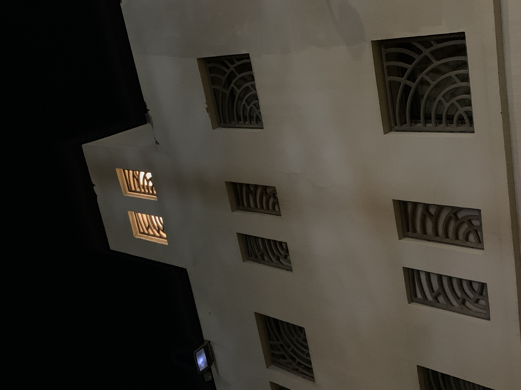 The image captures a nighttime view of a building facade from a low, upward-looking angle. The primary subject is a multi-story structure with a light-colored, possibly cream or beige, exterior. Most of the background is a deep, unlit black, indicating darkness or night sky.
Several rectangular windows are visible across the building's surface. A prominent feature of these windows is the elaborate, decorative metal grilles or screens that cover them. These grilles exhibit a repeating pattern of concentric, semi-circular or arc-like shapes. While most windows are dark, two on the left side of the building are brightly illuminated with a warm, yellow-orange light from within, suggesting interior activity or lighting.
On the lower left portion of the building, a small, square light fixture with a blue-white glow is attached to the wall. The overall scene is architectural, focusing on the patterns and contrasts of light and shadow on the building's exterior. There are no people visible, and no specific activity is taking place. The time of day is clearly night due to the dark sky and the reliance on artificial light from the building. No text is visible in the image. The architectural style, particularly the window grilles, could suggest a location in the Mediterranean or Middle East, aligning with the Tel-Aviv, Israel context.