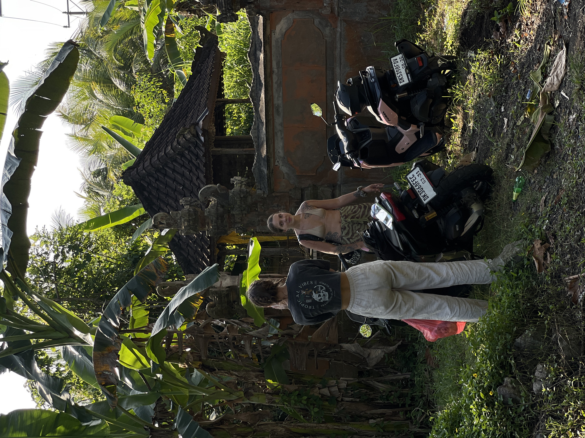 Two individuals, identified as females, are standing next to two scooters in what appears to be a rural area of Kerambitan, Indonesia. The individual on the left is wearing a dark long-sleeved crop top with graphics and light-colored linen pants. The individual on the right is wearing a light-colored bikini top and patterned shorts. Both are facing away from the camera, with the individual on the left appearing to be looking at something to their left, and the individual on the right looking towards the camera.
The environment features lush green vegetation, including large banana leaves dominating the left side of the frame. In the background, a traditional Balinese structure with a dark tiled roof is visible, suggesting a temple or a residential compound. The ground is covered in grass, dirt, and fallen leaves. One scooter is dark colored with a red accent, and the other is light pink. Both scooters have license plates. The scooter on the right has a plate that reads "DK5398FDV 08-2G". The scooter on the left has a plate that reads "DK3668FEC 03.31". A red plastic bag is visible near the legs of the individual on the left. The lighting suggests it is daytime, possibly late afternoon, with sunlight filtering through the leaves, creating shadows.
