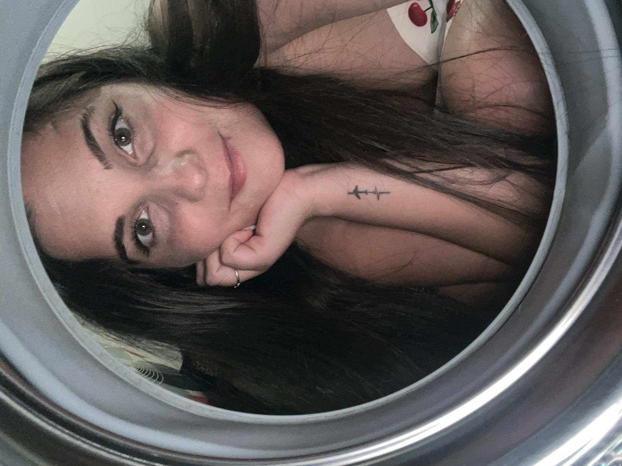 A woman with long, dark hair and brown eyes is looking into the camera. She has winged eyeliner and is resting her chin on her hand. She is wearing a ring on her finger and has a small tattoo of two airplanes on her forearm. Her clothing appears to be a white top with red cherries on it. The image is framed by a circular, metallic rim. The location is not specified in the image or accompanying information.
