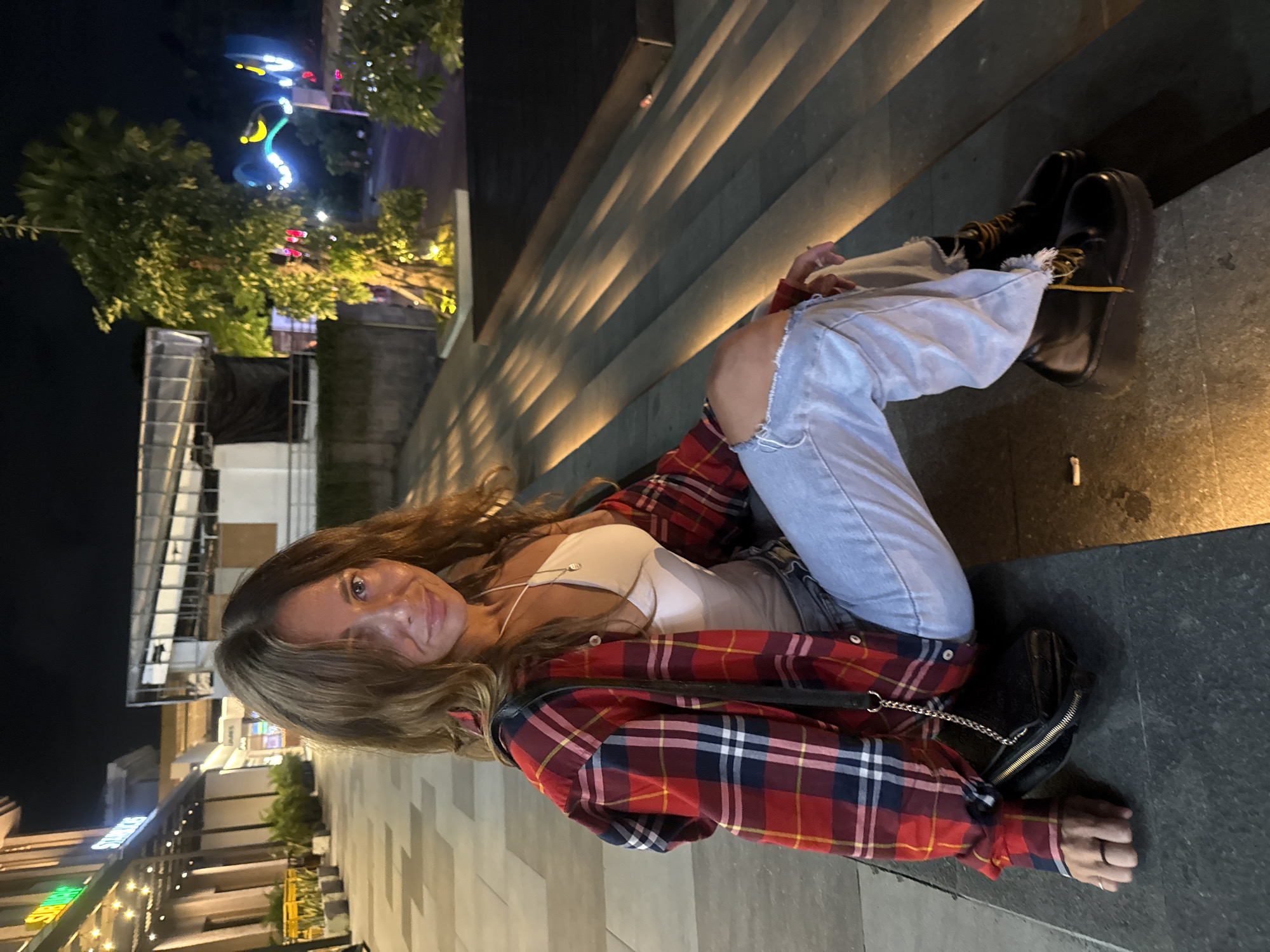 A woman sits on stone stairs in Kuta, Indonesia at night. She wears ripped light blue jeans, a white top, and a red and blue plaid shirt, along with black boots. She has long brown hair. The background shows buildings and trees illuminated by streetlights and decorative lights. The steps she sits on are also illuminated by strip lights.