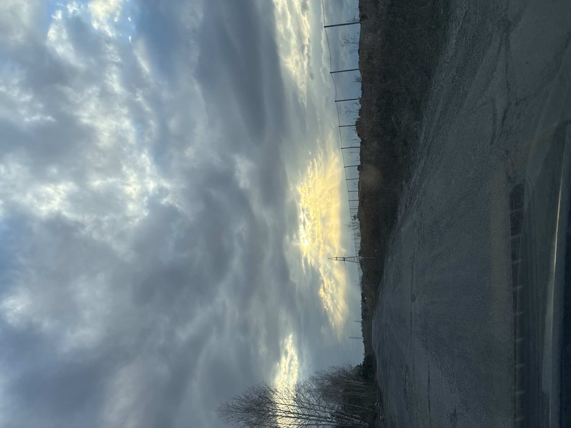 The image depicts a sky with striated clouds and a break in the cloud cover revealing sunlight. Below the sky is a landscape featuring a road, a grassy embankment, and a chain-link fence. There is an electrical transmission tower visible in the distance. Bare trees are present in the foreground. The location is Unknown, Unknown.