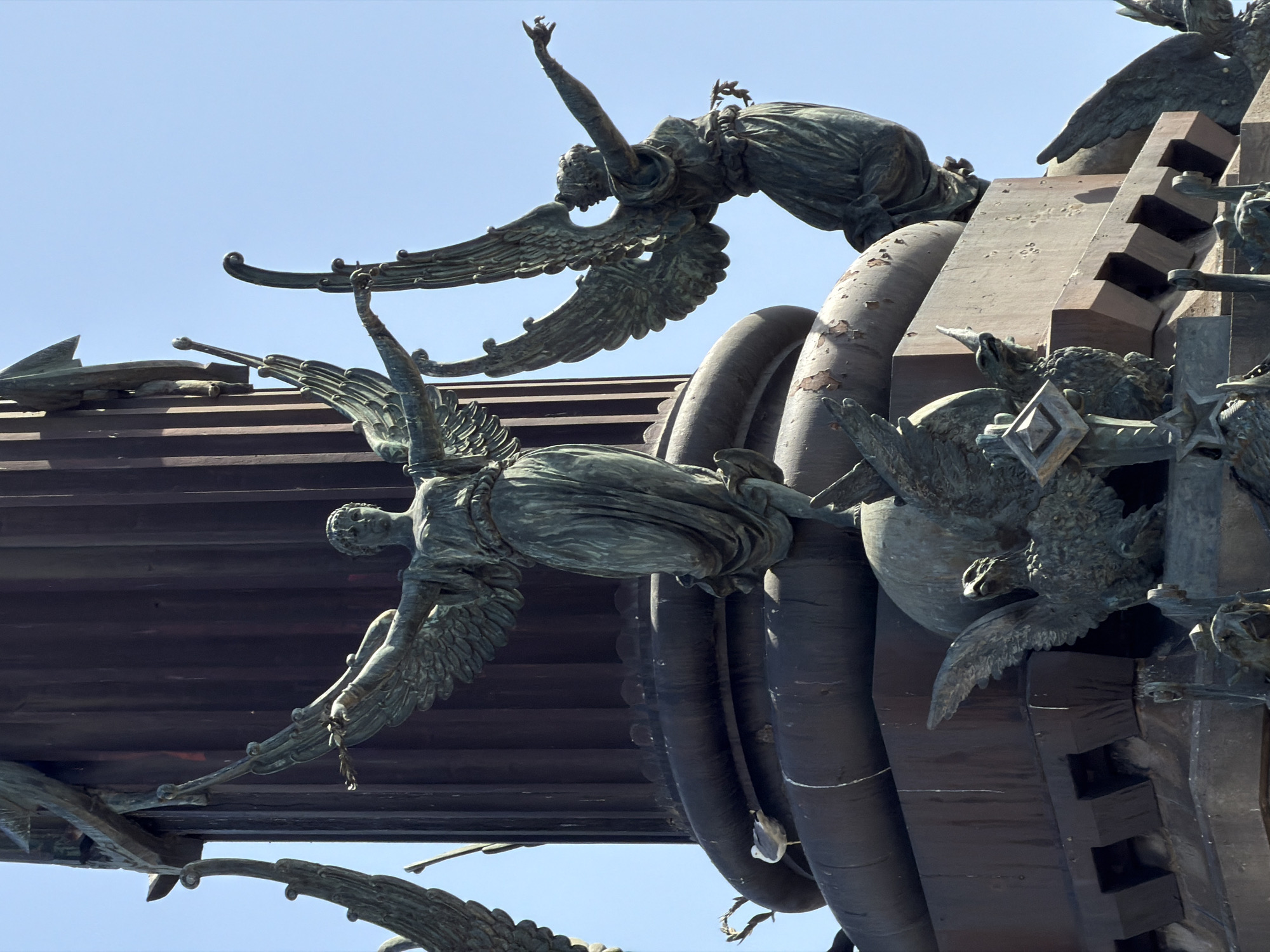 This image shows a detail of the Columbus Monument in Barcelona, Spain. The monument is made of bronze and depicts Christopher Columbus standing on a column, pointing towards the sea. The statue of Columbus is surrounded by allegorical figures and reliefs. In this specific shot, we see winged figures in classical attire, possibly representing victory or fame, with outstretched arms and wings spread. One of the winged figures is on the right, leaning forward with one arm extended. Below these figures, at the base of the monument, are depictions of eagles or griffins, with detailed feathers and beaks. A seagull is perched on a ledge of the monument in the lower left portion of the image. The monument is set against a clear blue sky. The bronze surfaces of the sculptures show signs of age and weathering, with some patination and slight discoloration.