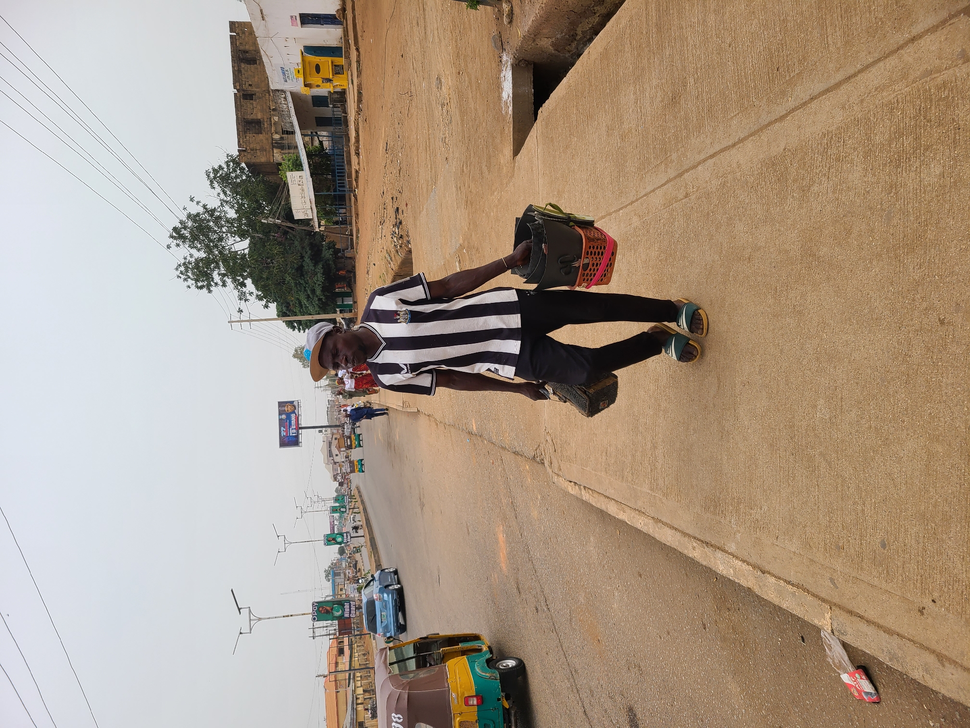The image displays a daytime street scene in an urban or semi-urban environment, likely in Jos, Nigeria, under an overcast sky. The central subject is a man of apparent African descent, walking along a raised concrete sidewalk towards the right side of the frame. He is dressed in a black and white striped short-sleeved shirt (resembling a football jersey), black trousers, light-colored sandals, and a light-colored hat that partially shades his face. In his left hand, he carries a dark-colored basket containing a smaller pink-rimmed basket, along with another dark bag.
To the man's left, a paved road stretches into the distance, bustling with activity. Vehicles, including a blue car and several multi-colored tricycles (known locally as Keke Napeps), are visible on the road. One tricycle has the partial number "88" displayed on its side. The road is lined with utility poles supporting overhead power lines and various billboards. Visible text on these billboards includes "I'M DRIVING", "OPay", and "NIGHT GUARD."
In the background, a mix of buildings with diverse architecture can be seen, some appearing older. One building features a sign partially reading "HOLLANDI." Green-leafed trees are scattered along the roadside. The concrete sidewalk has a visible textured surface, and a drainage channel runs alongside it. A discarded plastic wrapper with red detailing lies near the edge of the road. The overall scene depicts a typical street environment with ongoing daily life.