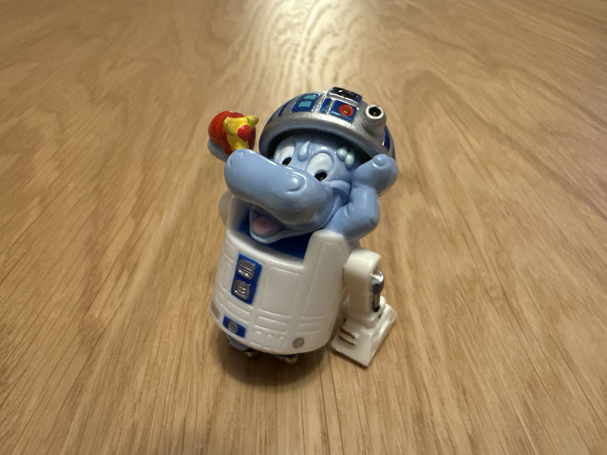 A toy figurine sits on a light brown wood-grained surface. The figurine is a blue hippopotamus wearing an R2-D2 themed helmet and body. The hippo is holding a red and yellow object in its left hand and has its right hand up to its head. The toy has R2-D2 design elements such as blue and silver panels on the helmet and white body with blue accents. The background is the wood surface. There are no indications of a specific location. Moscow, Russia is not mentioned.