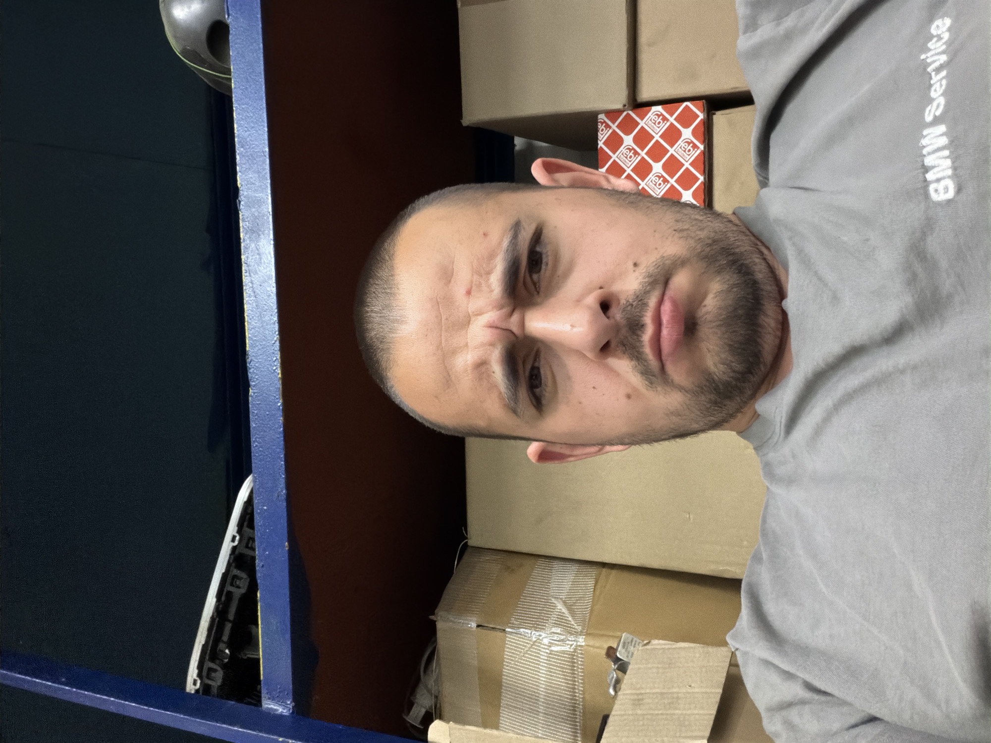 The image is a close-up, slightly rotated selfie of a man, taken indoors. The man, who appears to be in his 30s or 40s, has short, dark hair and a short beard and mustache. He is wearing a light grey long-sleeved shirt or sweatshirt with "BMW Service" embroidered in white text on the upper right side (when the image is oriented correctly). His eyebrows are furrowed, and there are visible wrinkles on his forehead, suggesting an expression of concentration or a slight scowl.
The background is dominated by stacked cardboard boxes, indicating an indoor setting like a storage room, workshop, or garage. Prominently visible near the man's head is a red and white box featuring the "febi" logo. To the left of the man, a dark blue metal frame or shelving unit is visible. On this frame, a dark, somewhat spherical object is partially visible, and further down, parts of a white circuit board or electronic component can be glimpsed through a gap in the structure. The overall lighting suggests an artificial indoor light source. The location is externally identified as Burgas, Bulgaria.