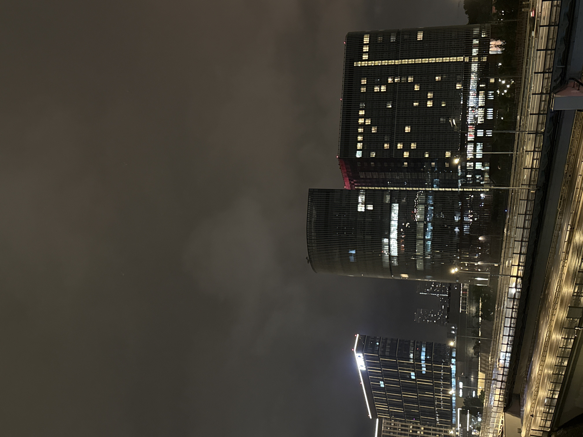 The image captures a night view of modern city buildings in Khimki, Russia, specifically showcasing the Novokosino skyscraper complex located near the MKAD highway. The sky is dark and cloudy, with the only significant illumination coming from the lights of the buildings and streetlights.
In the foreground, multiple levels of elevated highways are visible, with a few cars on them, indicating traffic flow. The roads are wet, suggesting recent rain. The highways are lined with railings and concrete barriers.
Dominating the mid-ground are three tall, modern buildings. The leftmost building is rectangular with a dark facade and vertical lighting elements, topped with illuminated white lines. The central building is cylindrical, featuring vertical ribbed detailing on its exterior and numerous illuminated windows. To its right is another rectangular building with a dark facade and a distinct red band near its top, also featuring many illuminated windows that create a grid-like pattern against the dark exterior. These buildings are part of the Novokosino skyscraper complex.
In the distance, behind the Novokosino buildings, other city lights are visible, hinting at a broader urban landscape. There are no visible people in the image. The overall scene depicts a well-lit urban environment at night, characterized by contemporary architecture and infrastructure.