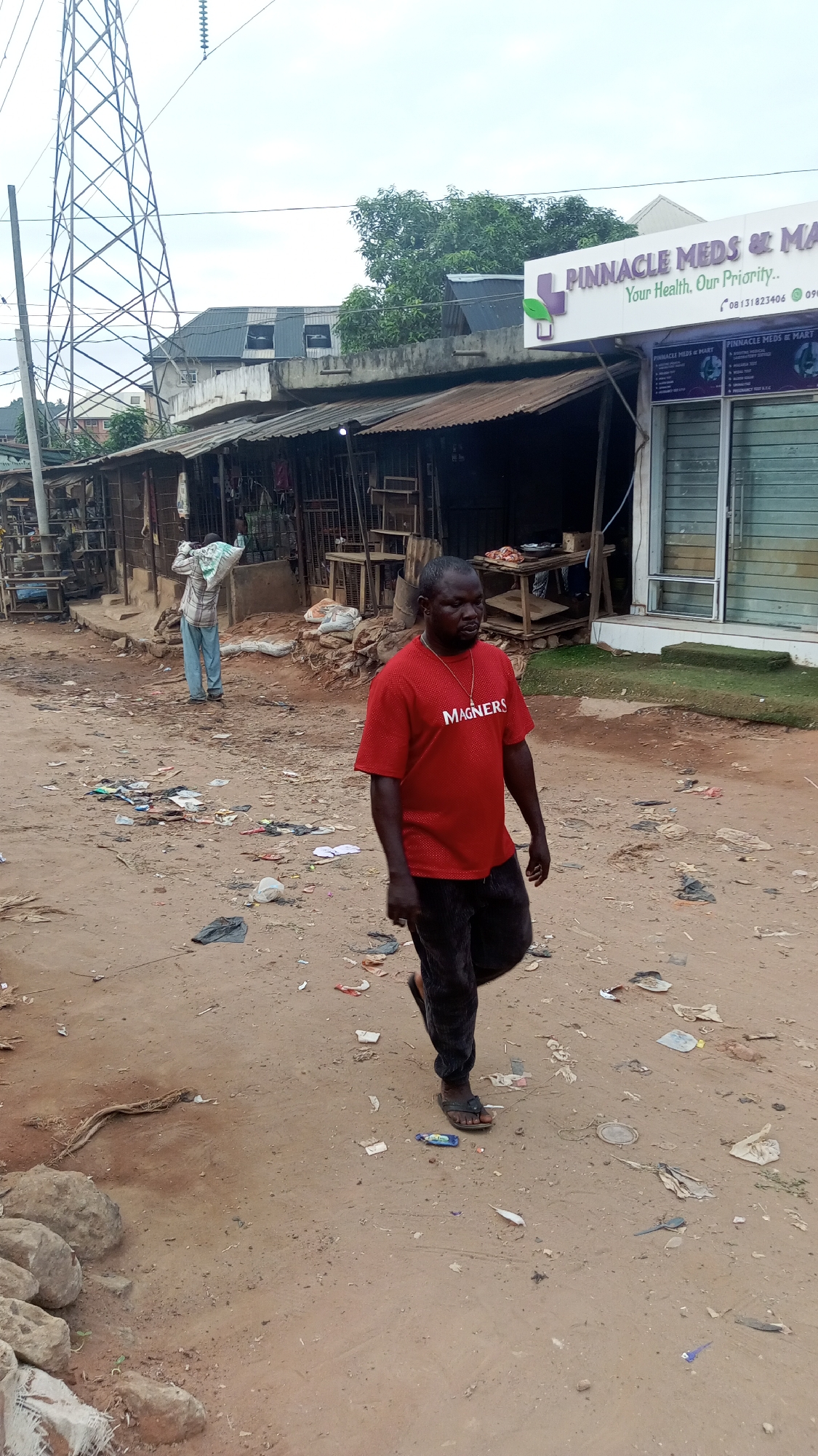 The scene is a dusty street in Onitsha, Nigeria. Debris litters the ground. A man wearing a red "MAGNERS" t-shirt and dark pants walks toward the camera, wearing sandals and a necklace. Another man with a sack on his back stands near a row of ramshackle stalls with corrugated metal roofs. Behind these stalls, a building with a sign reading "PINNACLE MEDS & MART" is visible. A tall, metal electrical tower stands in the background.