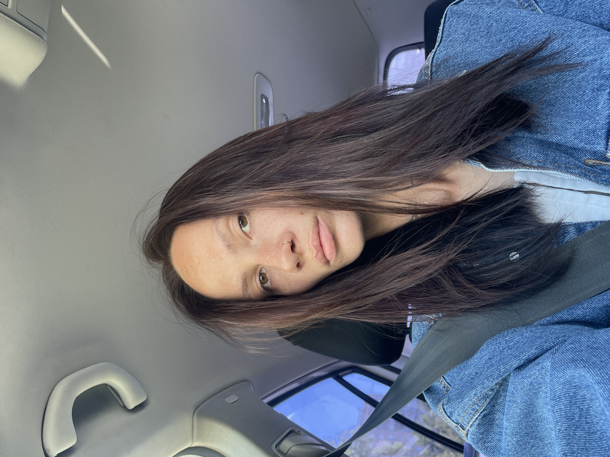 A woman with long, dark brown hair and fair skin is shown in a selfie taken from inside a car. She is wearing a blue denim jacket over a white top and her seatbelt is fastened across her chest. She has freckles on her face and a nose ring. The background shows the interior of a car, including the ceiling, a grab handle, a seatbelt, and a window with a blurred view of the outdoors. The image is taken during the daytime, with natural light illuminating the scene. There is no indication of the specific location in Izhevsk, Russia.