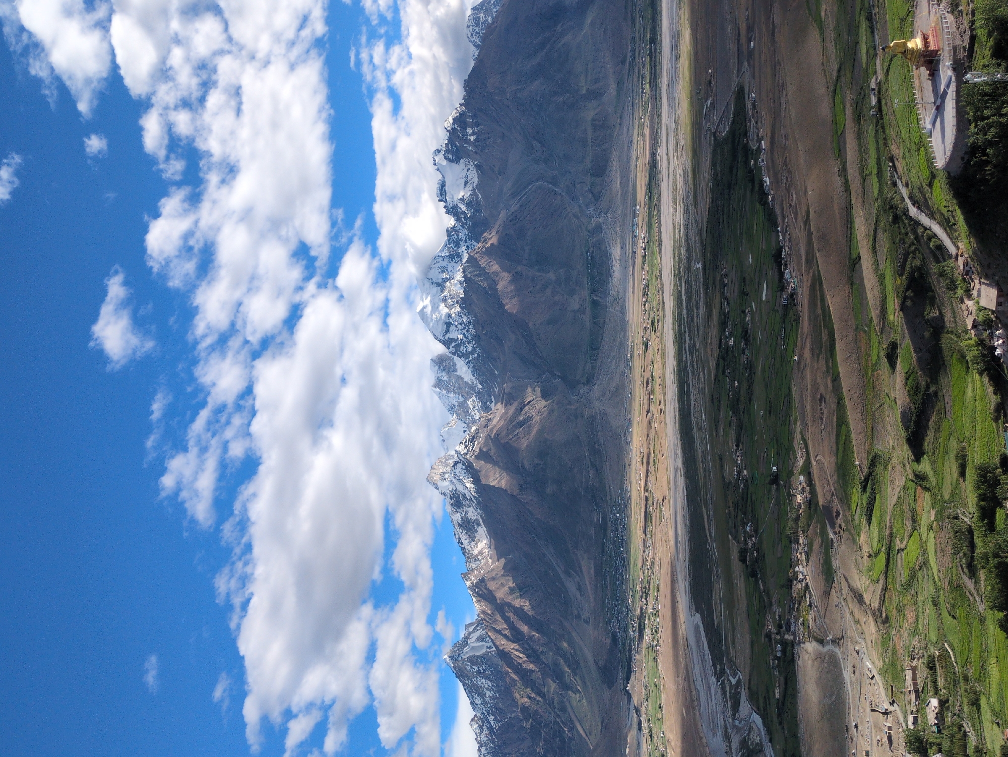 This is a vertical, high-angle, outdoor shot of the Yulang valley in Ladakh, India. The scene is dominated by a dramatic mountain range in the background, its peaks capped with snow and the slopes a mix of brown and grey. Below the mountains, the Yulang valley unfolds, a patchwork of green fields and brown, arid land bisected by a winding river. Small clusters of buildings, likely a village, are scattered across the valley floor. In the foreground, a golden Buddha statue sits atop a circular platform, a prominent landmark against the lush green slopes. The sky above is a vibrant blue, dotted with fluffy white clouds. The lighting suggests it is daytime, with shadows indicating the sun is to the left.