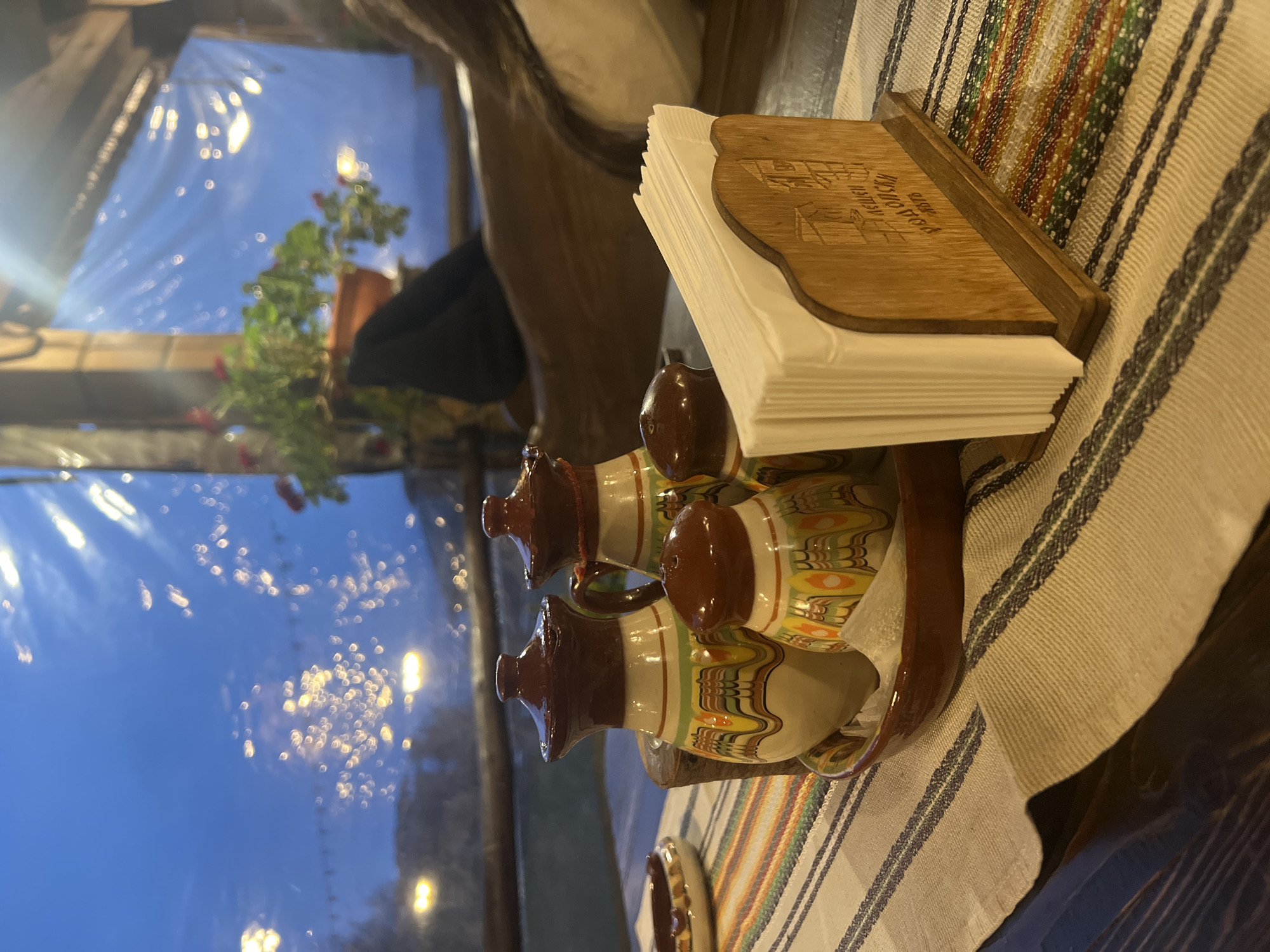The image shows a table setting in what appears to be a restaurant or cafe in Leshten, Bulgaria.
The table is covered with a striped woven cloth. On the table, there's a napkin holder containing a stack of white napkins, and a set of salt and pepper shakers with a traditional Bulgarian design. A glimpse of a potted plant and blurred lights is visible through the window in the background.