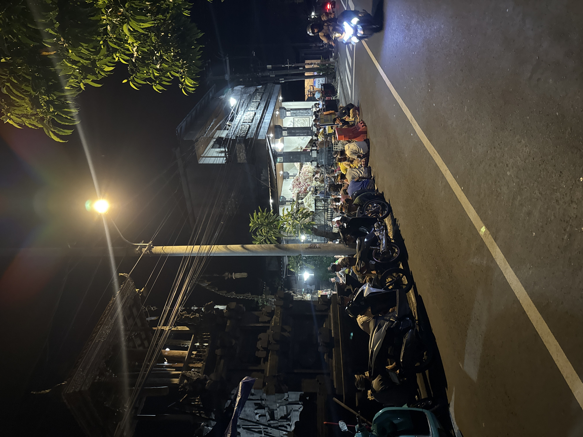 In Ubud District, Indonesia, at night, a group of people are gathered on the side of a paved road. They are sitting on the ground next to several parked motorcycles. Across the street, a multi-story building is illuminated, with more people visible inside and around its entrance. A motorcycle rider is actively moving along the road, their headlight on. The scene is lit by a street lamp, which casts a warm glow and creates lens flares. Trees with green leaves are visible on the right side of the frame, with their branches extending towards the street. The architecture of the building across the street appears to be traditional, with ornate carvings and dark wood accents.