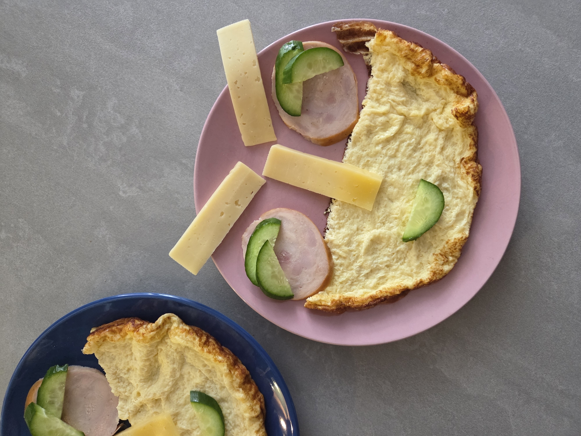 An overhead shot captures two plates of breakfast food on a gray, textured countertop, likely in a kitchen or dining area in Birobidzhan, Russia.
The primary focus is on a pink round plate in the upper right, laden with a substantial portion of a fluffy, golden-yellow omelet. The omelet has a slightly crisped edge. Alongside it are three rectangular slices of cheese, varying slightly in shade from light to medium yellow. Two thin, round slices of what appears to be ham or other cold cuts are arranged on the plate, with two fresh cucumber slices artfully placed on top of one of the ham slices. A single cucumber slice rests near the omelet's edge.
Partially visible in the lower left is a blue round plate, also holding a portion of the same type of omelet. On this plate, a slice of ham and two cucumber slices are visible. A hand, partially out of frame, holds another rectangular slice of light yellow cheese above the blue plate, as if about to add it to the meal.
The scene suggests a prepared breakfast or brunch, ready to be eaten. The lighting is bright and even, indicative of an indoor setting during daytime. There is no visible text in the image.