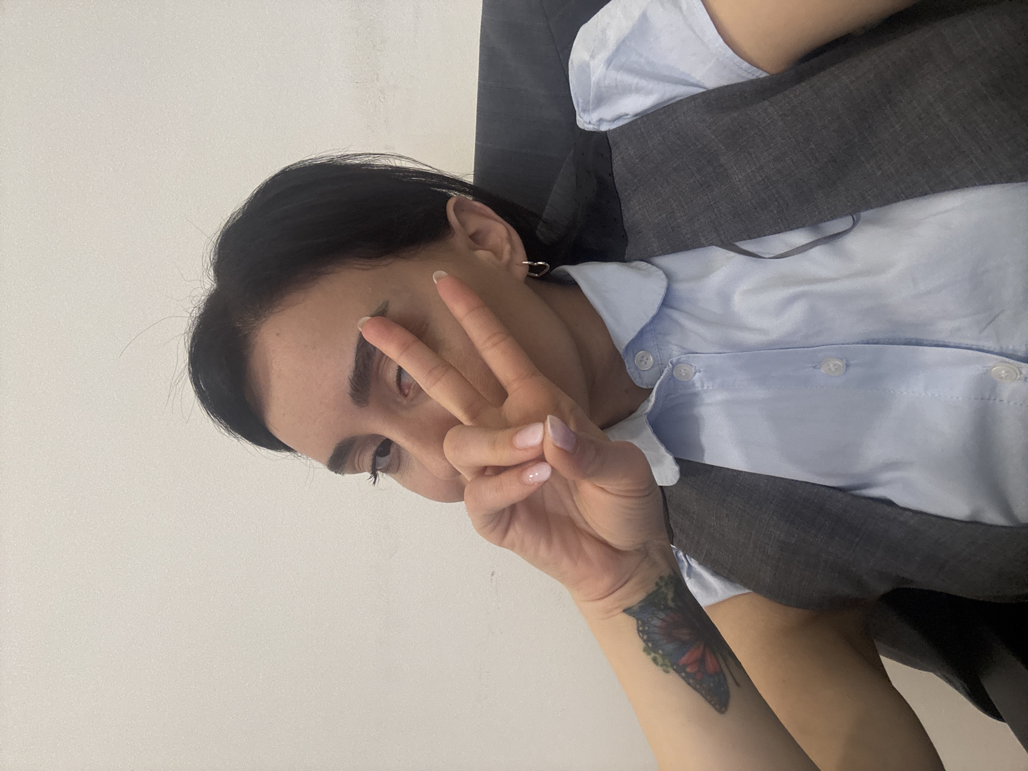 A woman with dark hair is pictured indoors against a light wall. She is wearing a light blue, button-down shirt and a gray vest. Her left hand is raised, making a peace sign near her face. A butterfly tattoo is visible on her left forearm. The background is a plain white wall. The image orientation is vertical. There is no indication of the location being Tbilisi, Georgia.
