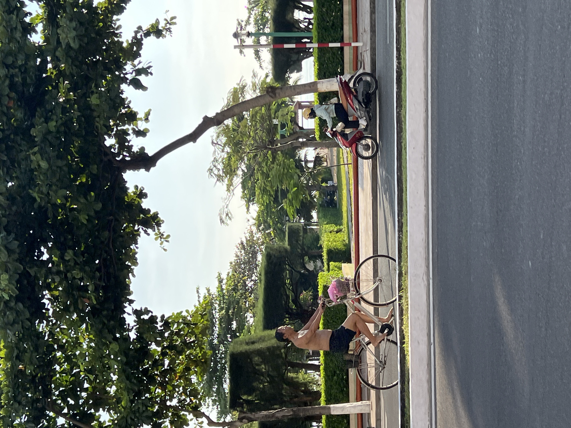 In Nha Trang Ward, Vietnam, a man without a shirt and wearing black shorts cycles on the left side of a paved path. He is barefoot. A pink bag is attached to the handlebars of his bicycle. To his right, a person wearing a light blue jacket and a yellow helmet rides a red scooter. The scooter rider is wearing white socks and dark pants. They are both on a road with a white and red striped pole to their right. The path is bordered by manicured green hedges and trees. The background features more trees and a cloudy sky.