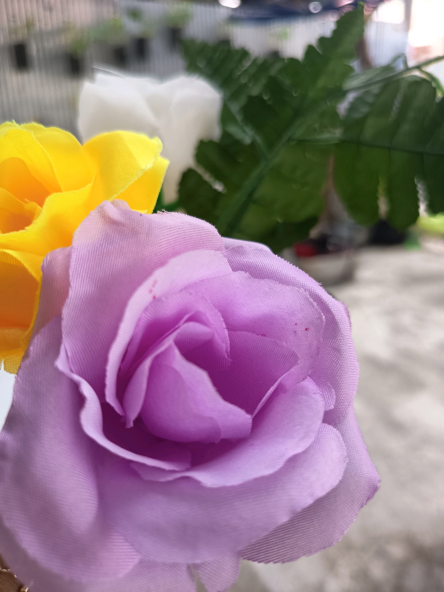 The image shows a close-up of three artificial flowers: a purple rose in the foreground, a yellow rose to its left, and a white rose behind the yellow one. A blurred green leaf is visible behind the white rose. The flowers appear to be made of fabric. The background is out of focus, showing a light-colored, textured surface and hints of green foliage in the distance. It is not possible to determine the exact location within Banjarmasin, Indonesia from this image alone.