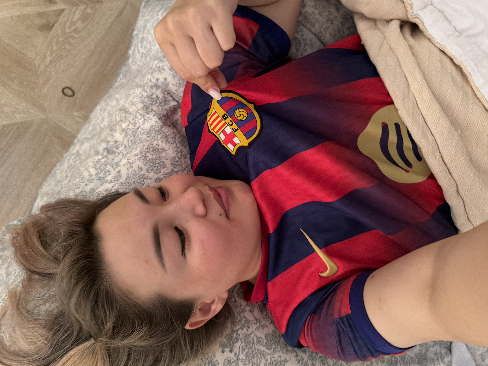 In this overhead shot, a young woman lies on a bed covered with a patterned gray duvet. She is wearing a red and blue striped t-shirt, possibly a sports jersey, with a crest on the left chest. Her right hand is shown touching the crest. Her eyes are closed, and she appears relaxed, with her head tilted to the side. Her hair is a light brown and styled loosely. The lighting suggests it is daytime, and the setting is indoors, likely a bedroom. There is no visible text in the image. The background shows a light-colored, wood-patterned surface on the left and a beige blanket on the right.