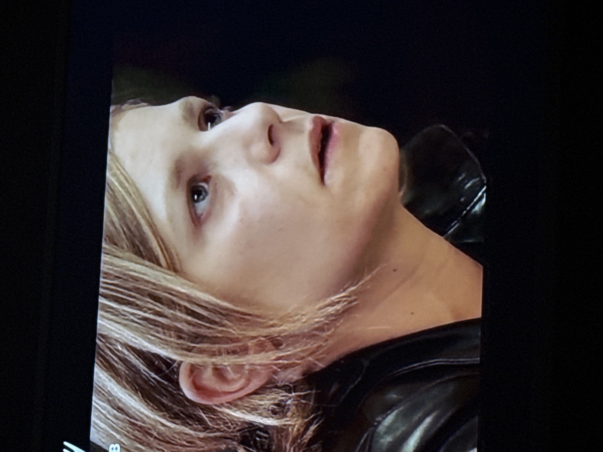 A close-up shot of a woman with blonde hair looking upwards. She is wearing a black leather jacket. The background is black. There is no indication of the location being Mytishchi, Russia.