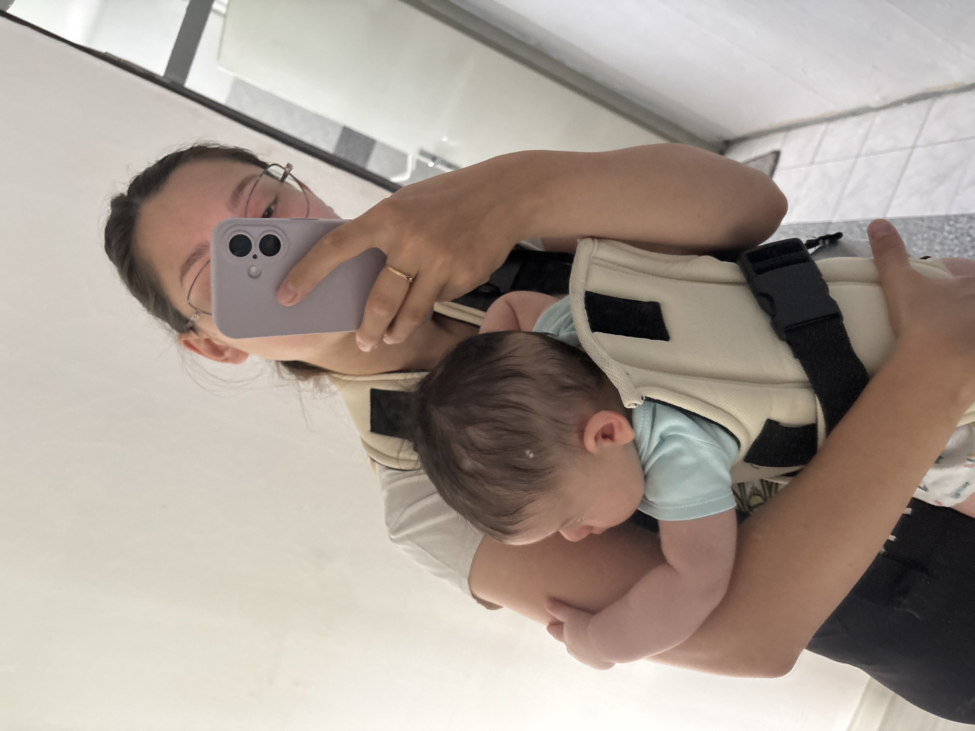 A young woman wearing glasses and a light-colored t-shirt is reflected in a mirror, holding a baby in a carrier. She is also holding a smartphone in her right hand, as if taking a selfie. The baby is wearing a light blue onesie and is facing away from the camera. The background appears to be a bathroom with tiled walls and a white wall. The scene is indoors. There is no information about the location being in Plovdiv, Bulgaria.