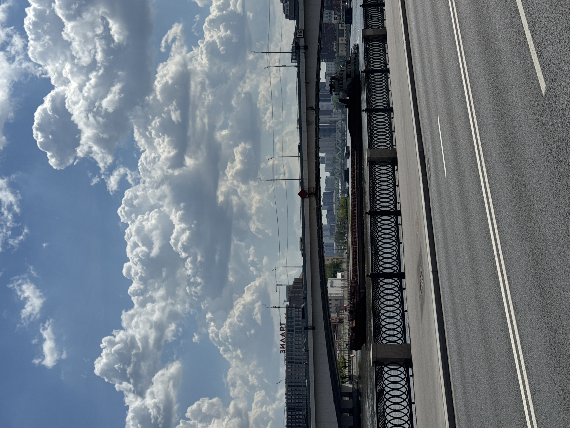The image, when correctly oriented, presents an elevated view from a bridge over a waterway and cityscape, under a bright, cloudy sky.
On the left side, a multi-lane asphalt road is visible, marked with white lines for traffic. It is bordered on its right by a decorative black metal railing, beyond which lies the concrete structure of the bridge. Several streetlights and utility lines run along the bridge's length.
Below and beyond the bridge, a wide waterway, likely a river, stretches towards the horizon. Along its far bank, a dense urban landscape unfolds, characterized by numerous high-rise buildings and other architectural structures. A prominent tall building in the middle distance clearly displays the Cyrillic text "ЗИЛААРТ" (ZILART) in red letters. On the bridge structure itself, a sign with the text "Б1Б" is visible. Several boats, including what appears to be a large barge or ferry, are present on the water near the far bank.
The sky above is a mix of bright blue patches and large, white, voluminous cumulus clouds, suggesting fair but dynamic weather conditions. The scene is illuminated by natural daylight, indicating a time between late morning and afternoon. No people are visible in the frame. The presence of the "ZILART" signage, coupled with the provided context, confirms the location as Moscow, Russia.