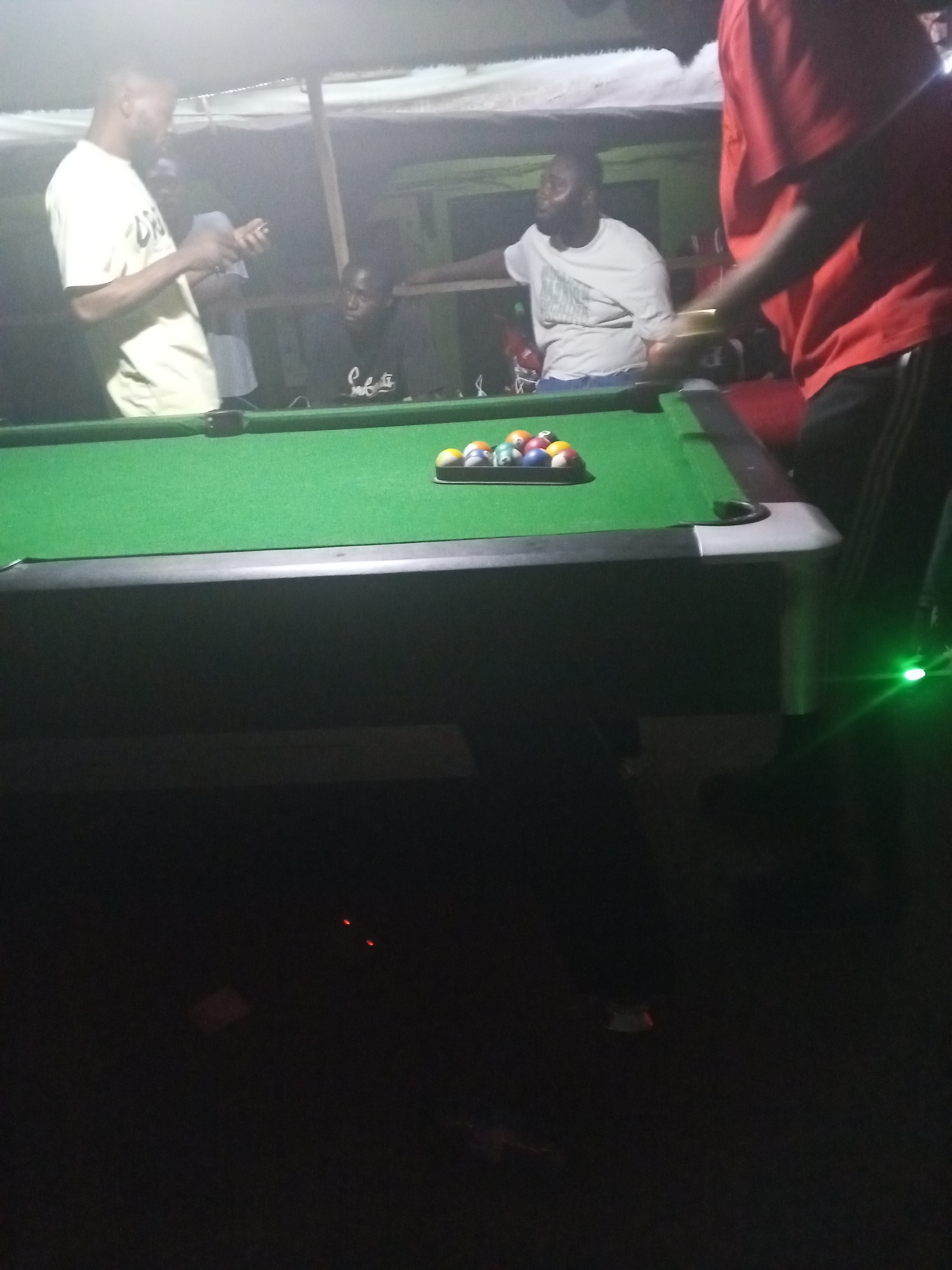 The image captures a dimly lit, casual gathering around a pool table, likely at night, in Lokoja, Nigeria. Four men are visible in the scene. To the far left, a man in a light-colored t-shirt with faint text, possibly "DREAM," is standing and looking down at a smartphone he holds in both hands. Behind the pool table, a man in a black t-shirt with "Swifte" or similar text is seated or leaning, looking towards the left side of the frame. Further to the right, another man in a white t-shirt with some indiscernible text is leaning on the pool table's edge. A fourth man, partially visible on the far right, is wearing a red t-shirt and dark pants with white stripes, standing near the table.
The central object is a green-felted pool table. A rack of billiard balls, comprising both solid and striped balls, is neatly arranged near the center of the table. The table itself has dark grey or black siding. Above the group, a makeshift shelter made of corrugated iron sheets supported by wooden beams is visible. The overall lighting is dim, suggesting evening or nighttime, with artificial light sources illuminating the top portion of the scene. A bright green light or laser dot is visible on the lower right side, near the base of the pool table. The atmosphere appears relaxed and social.