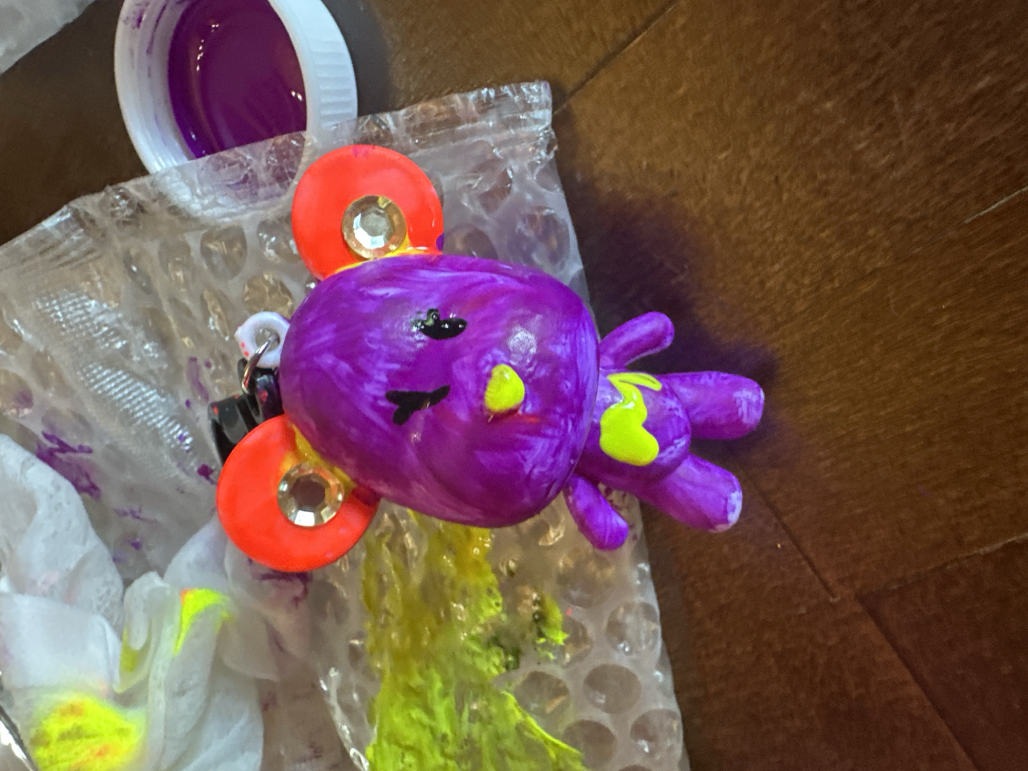 A high-angle shot depicts a crafting scene on what appears to be a wooden surface. A purple, painted clay figure resembling an octopus is the central subject, with lime green paint detailing on its tentacles and head. Two orange circular pieces with embedded rhinestones are attached to the octopus' head. A jar containing purple paint is positioned on the top left of the frame. A sheet of bubble wrap covers much of the scene, with splatters of yellow paint on it and on a white cloth.