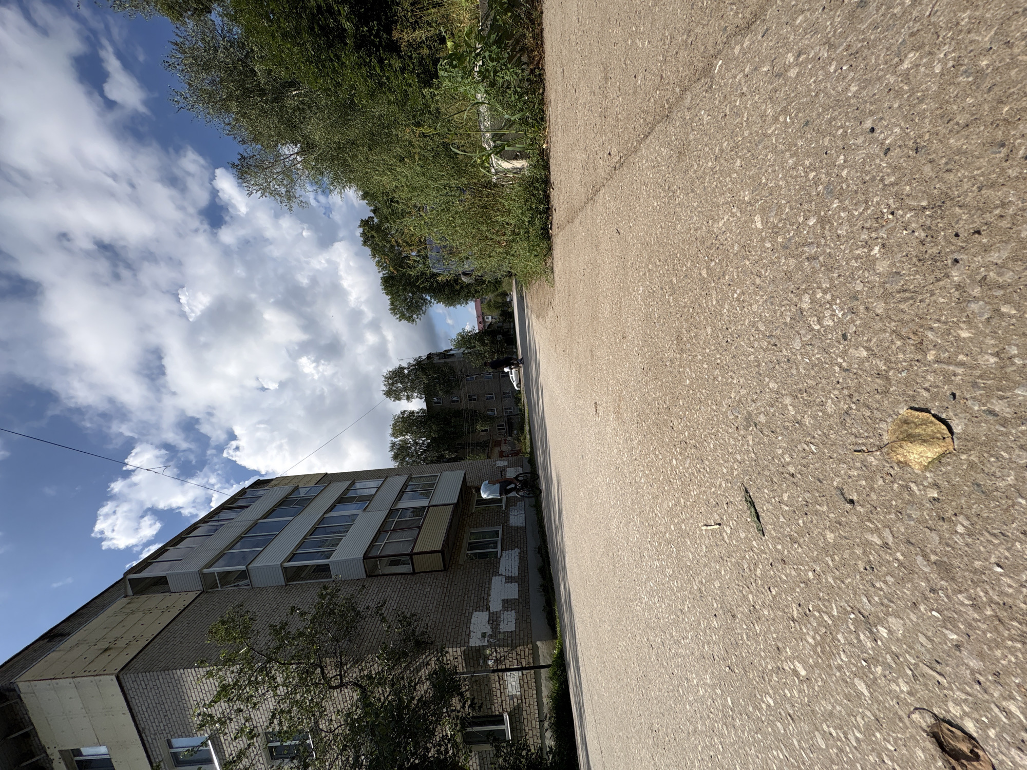 This is a low-angle, wide-shot photograph taken on a sunny day in the city of Krasnokamsk, Russia. The foreground is dominated by a paved asphalt surface with scattered leaves and debris. To the left, a multi-story brick apartment building is visible, with some signs of wear and tear, including patches of white on the lower facade. A tree with green foliage grows near the building. In the middle ground, a person on a bicycle is riding away from the camera. Further down the street, more apartment buildings and dense green trees line the road. The sky is blue with scattered white clouds. The scene depicts a typical urban residential area.
