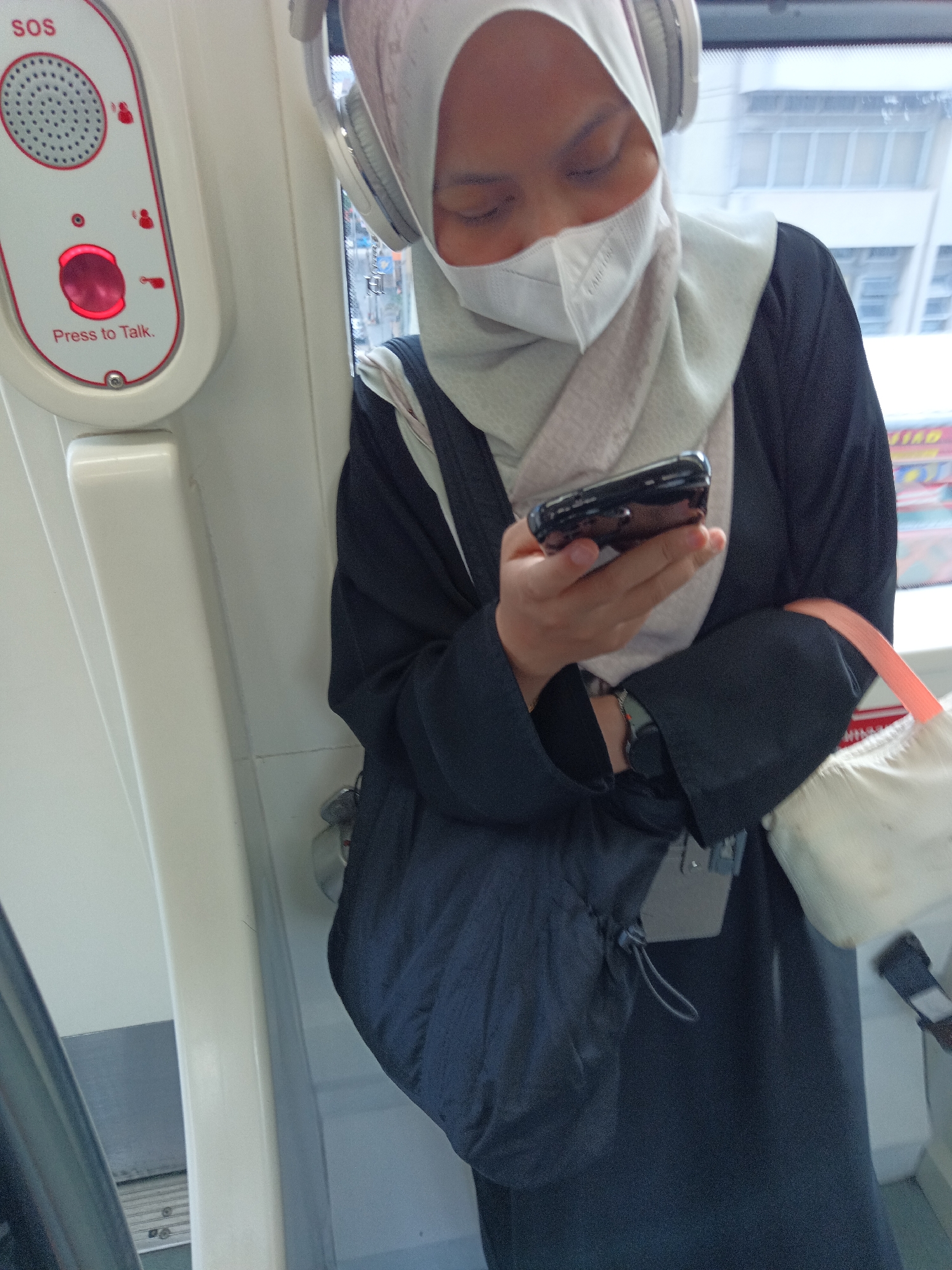 A person wearing a white hijab, a white face mask, and a dark colored abaya is standing inside a vehicle, likely a train or bus, and is looking down at their phone. They are also wearing headphones. To the left of the person, there is a control panel with an "SOS" label and a red button that says "Press to Talk." The individual is holding a black smartphone in their hands. A light-colored bag with an orange strap is slung over their shoulder. The background shows a blurred cityscape with buildings and some signage, suggesting an urban environment. Given the presence of public transportation with an SOS button and the general aesthetic, it is plausible this scene is within the public transport system of Kuala Lumpur, Malaysia, such as the LRT or MRT.